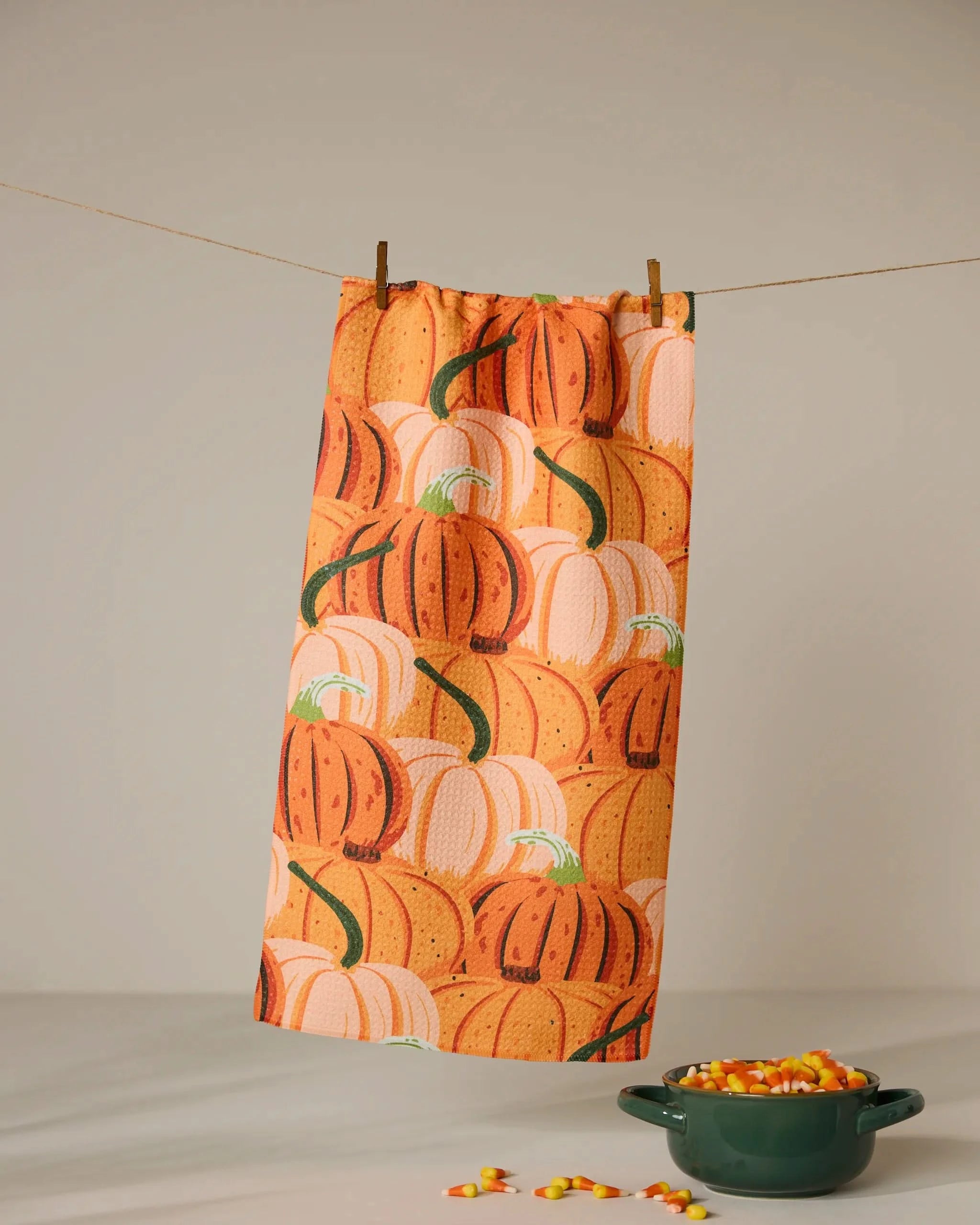 The Geometry Rows of Gourds bar towel, featuring an orange pumpkin pattern, hangs from a clothesline with clothespins. Below, a green bowl of candy corn adds to the festive display. This ultra-absorbent towel is both stylish and practical.