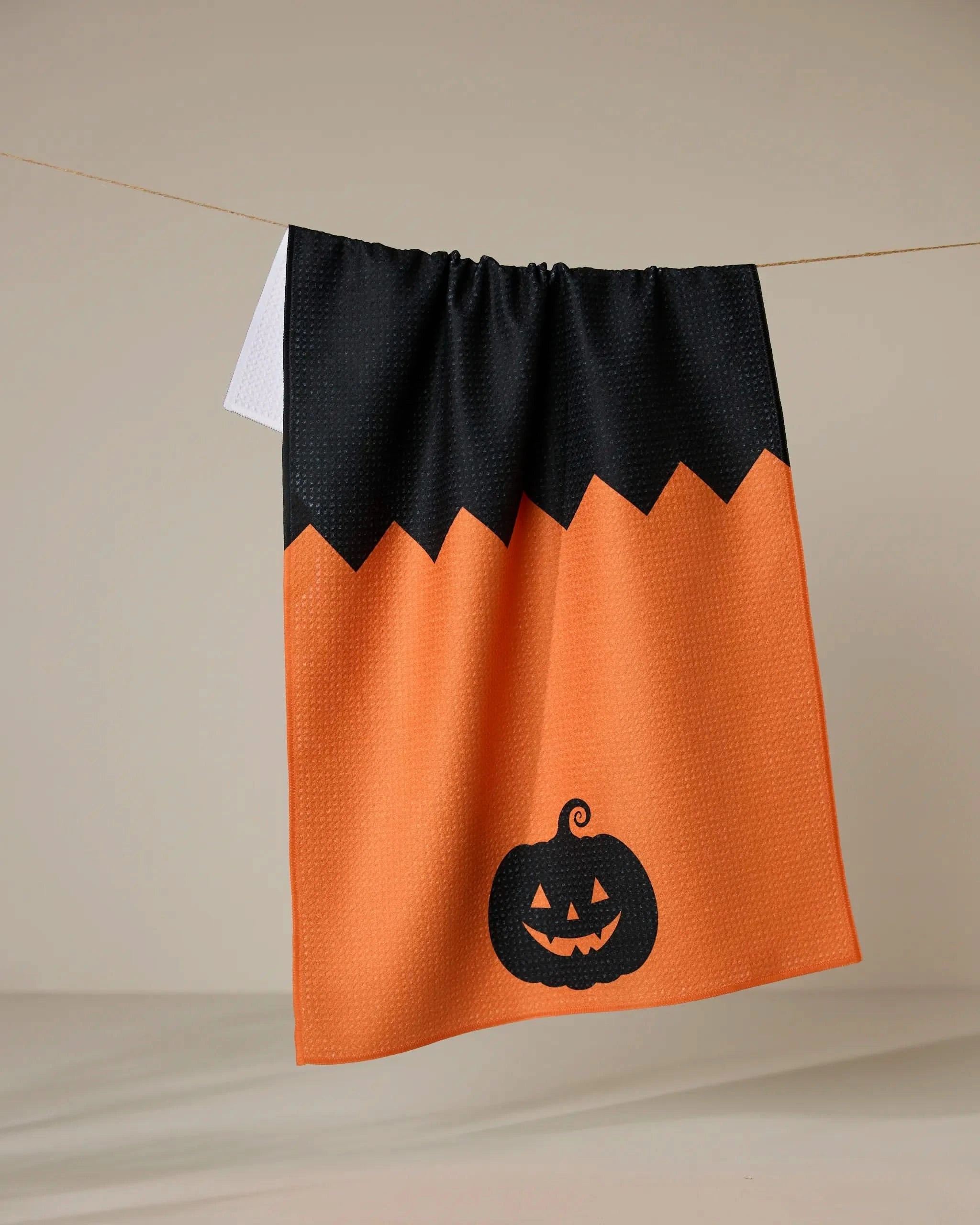 The Geometry Spooky Split tea towel hangs on a line. Made from recycled material, this orange towel features a black zigzag border and a black jack-o-lantern design near the bottom.