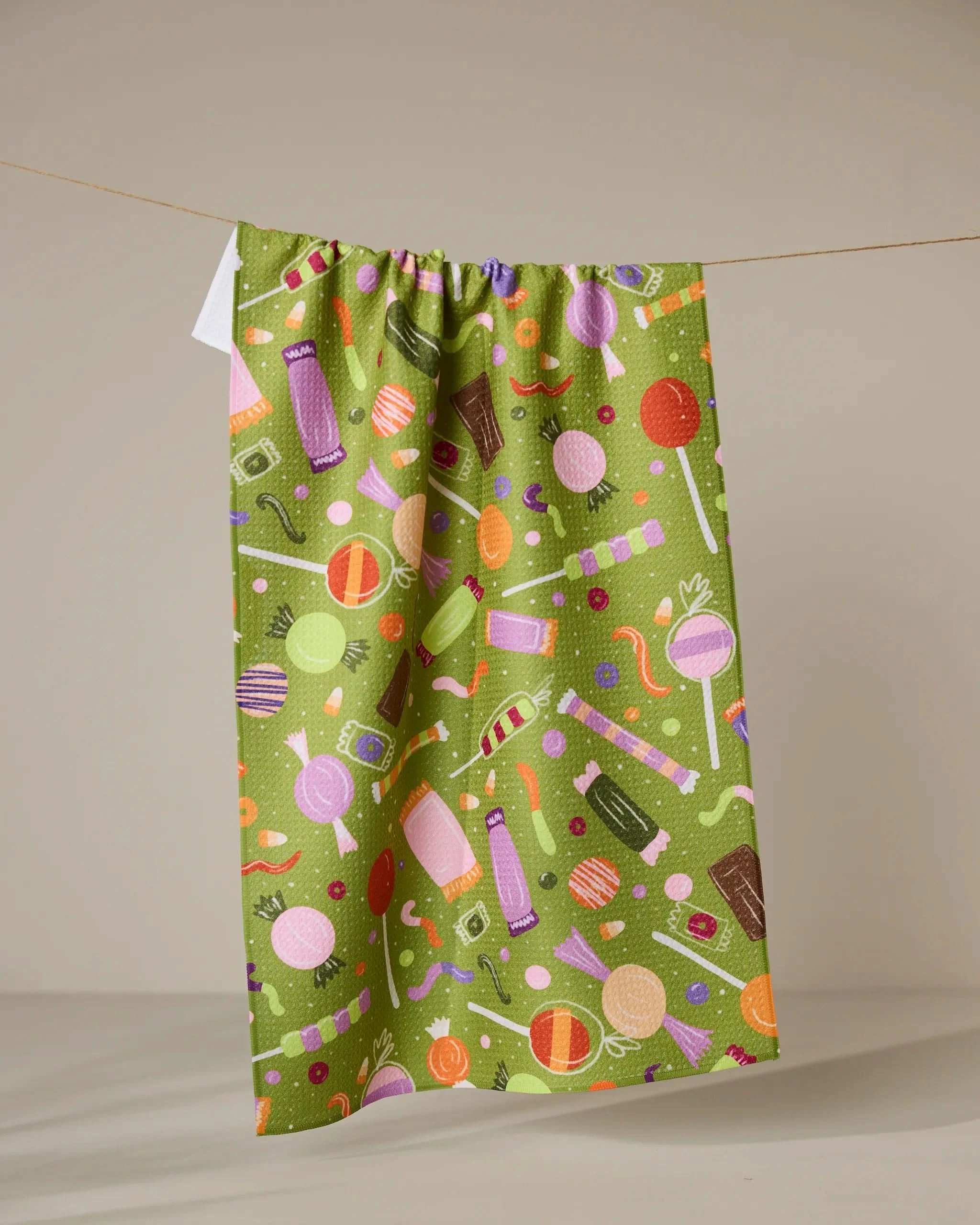 Sweet Treasures by Geometry: A green tea towel with vibrant candy graphics—lollipops, wrapped sweets, chocolate bars, and gummy worms—hangs against a beige background. Made from ultra-absorbent recycled materials.