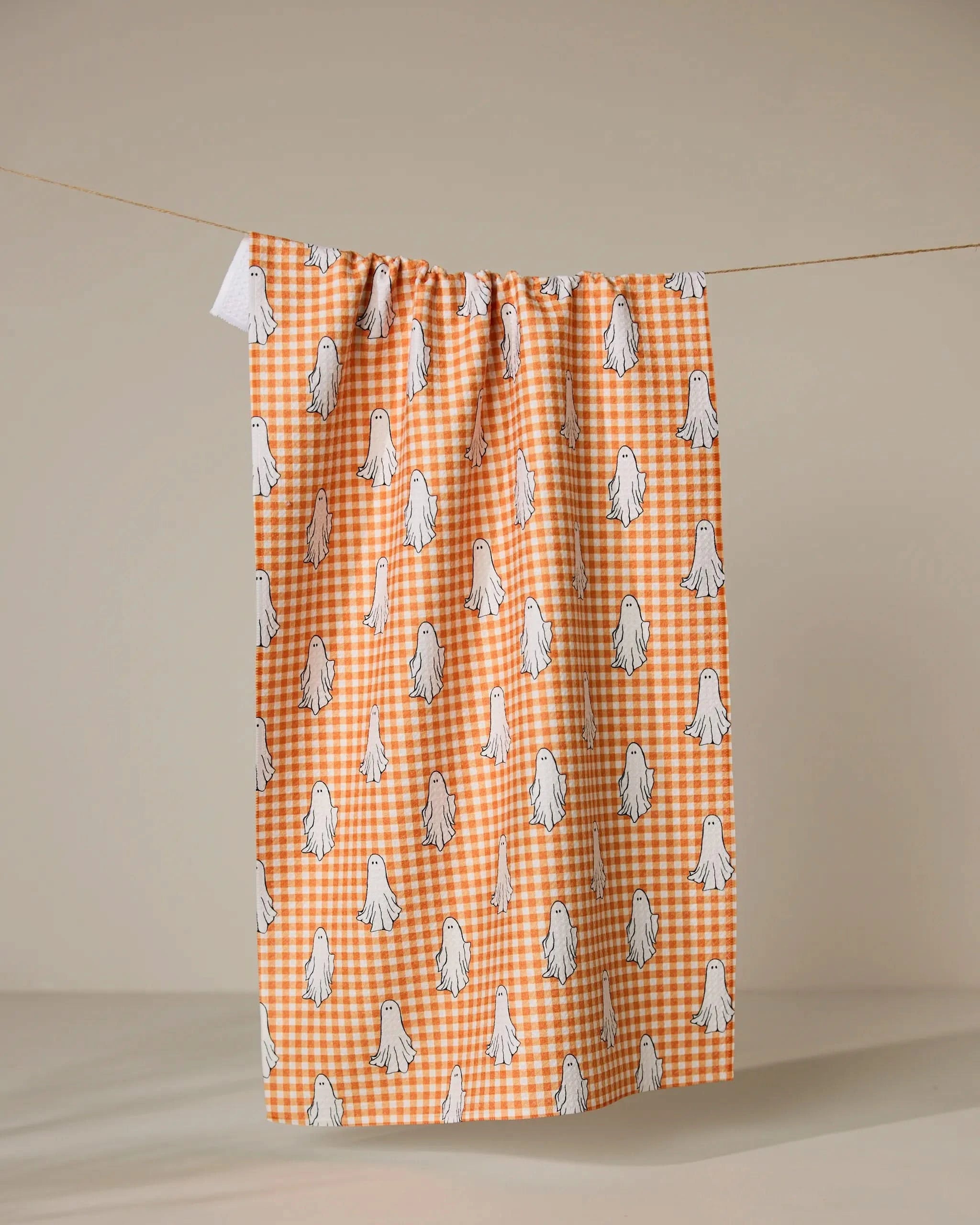 The Geometry Gingham Ghosts tea towel, crafted from recycled materials, features an orange and white gingham design with a cute repeating pattern of small white cartoon ghosts.