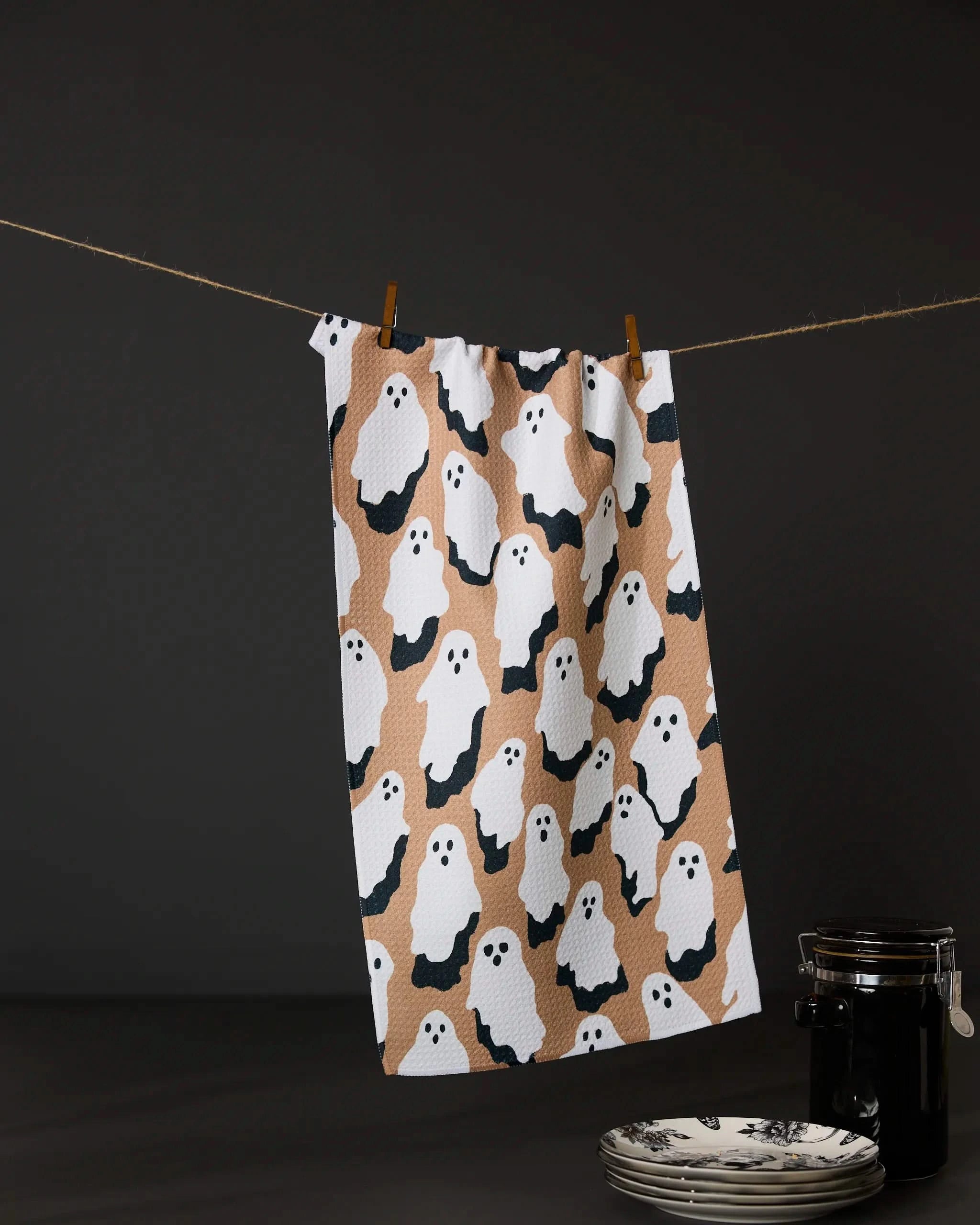 The Geometry My Boo recycled kitchen towel, featuring white cartoon ghosts on a brown and black background, hangs from a clothesline above stacked plates and a black jar against a dark backdrop.