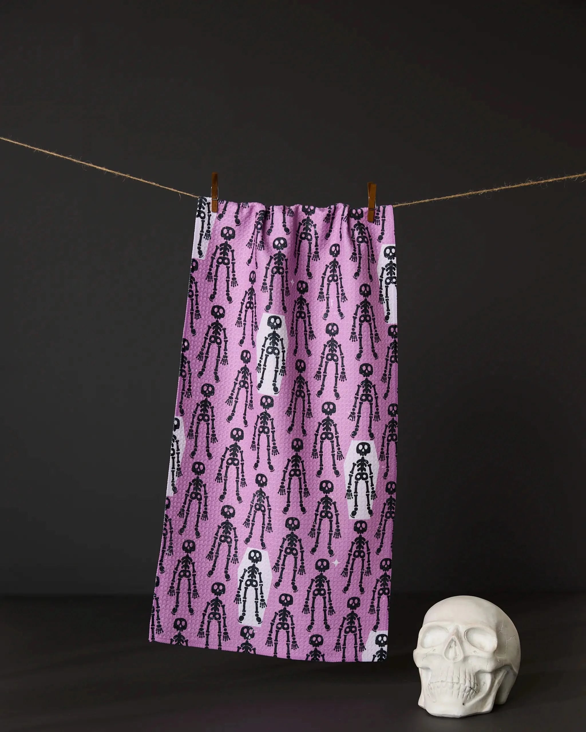 The Geometry Bones and Beds towel, featuring a black and white skeleton pattern on pink, hangs on a clothesline above a white skull decor with a dark background.