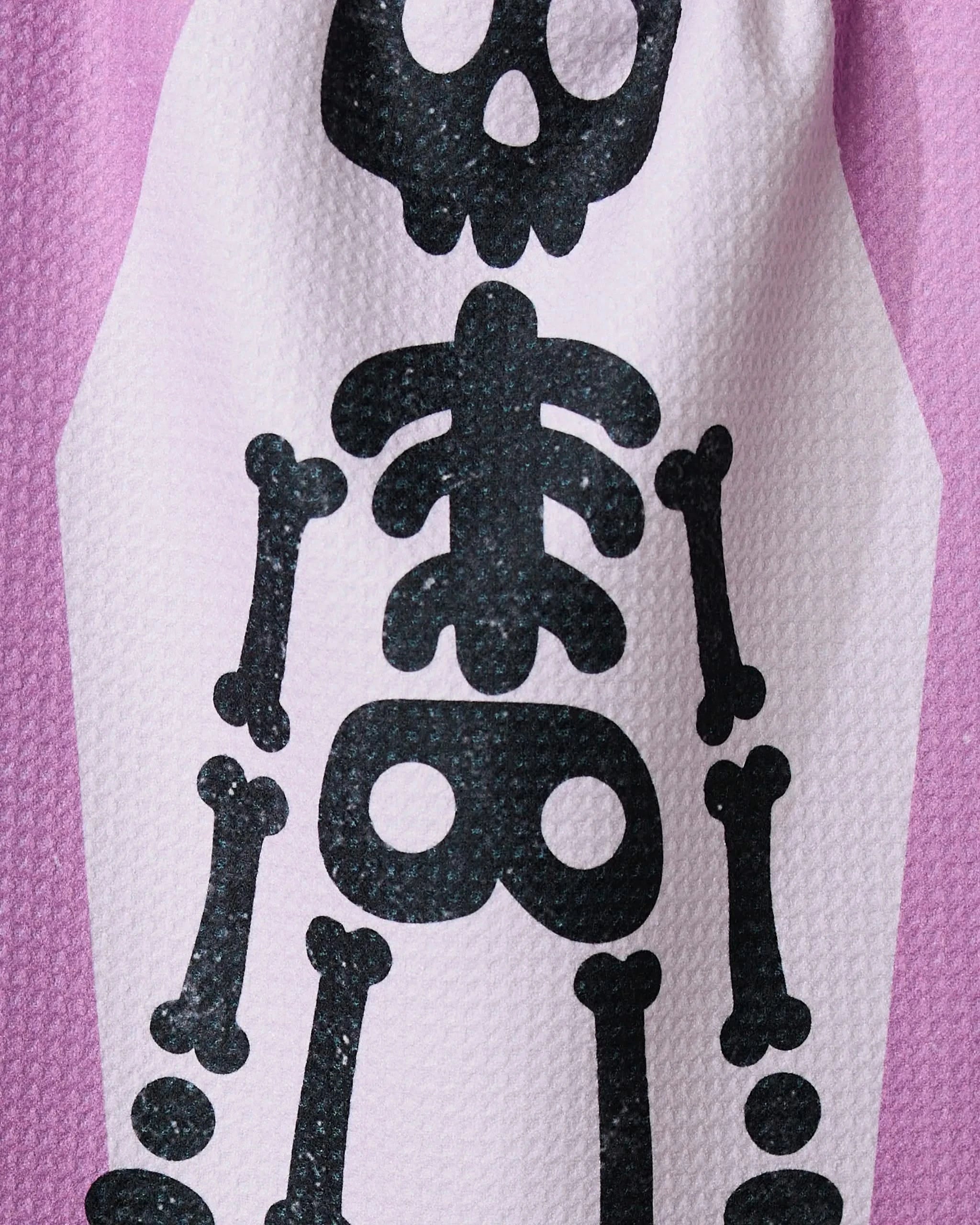 A close-up of Geometry’s Bright Bones, an ultra-absorbent towel made from recycled materials, features a black cartoon skeleton print with skull, ribcage, and upper leg bones on light pink textured fabric.