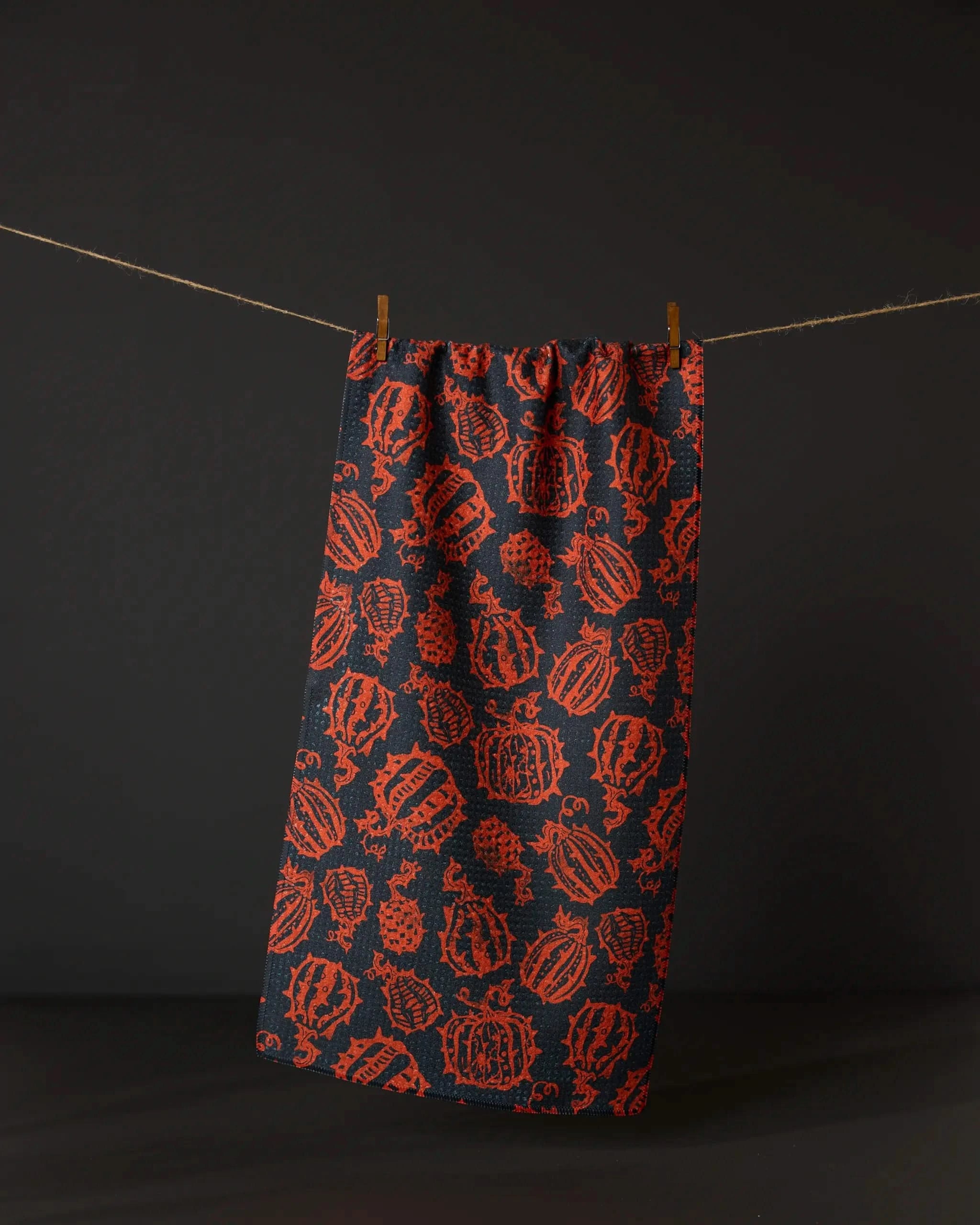 Geometrys Punk Pumpkins Black bar towel, featuring a red repeating pumpkin pattern, hangs by two small clothespins on a string against a black background.