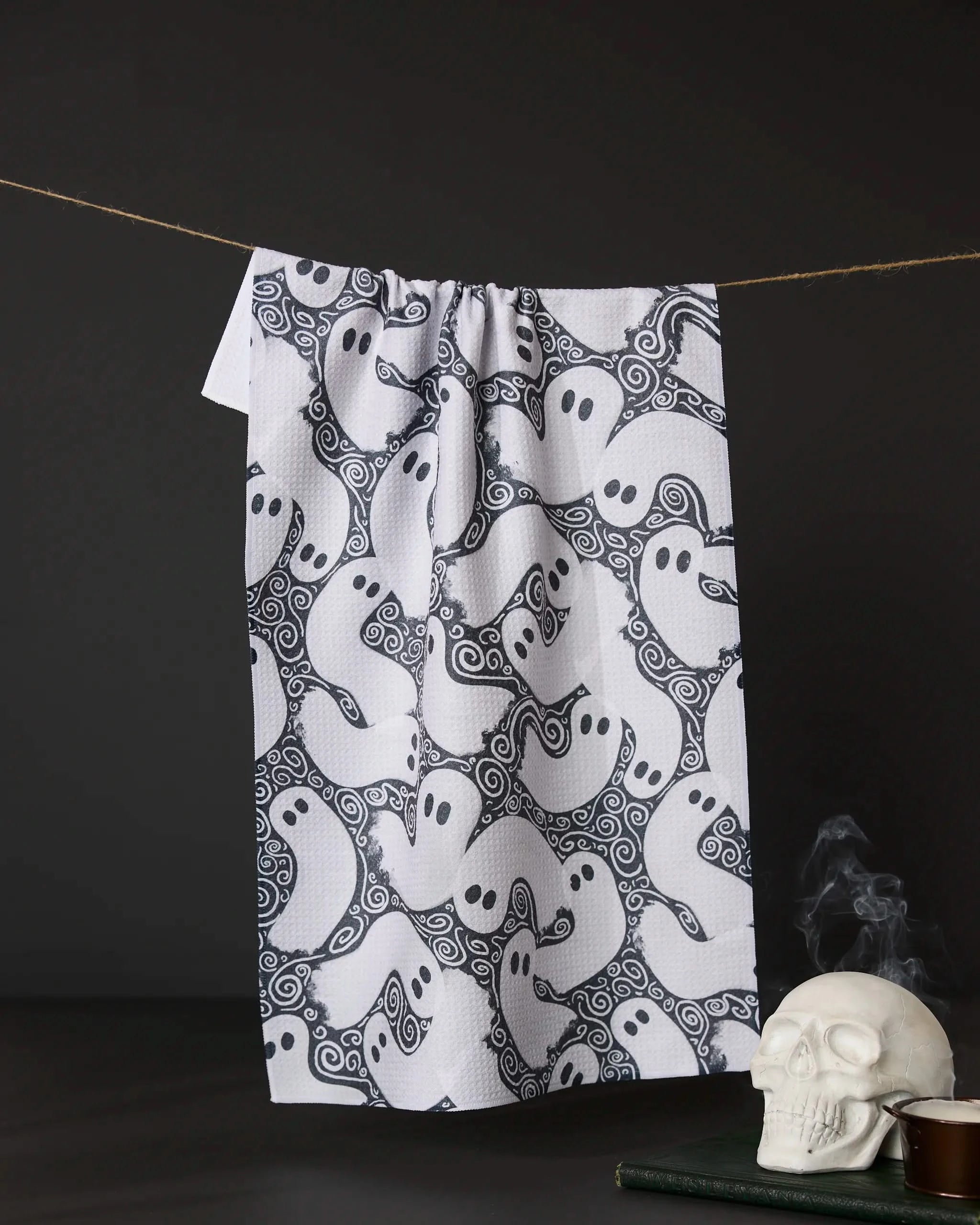 The Geometry Swirling Ghosts tea towel, featuring a cartoon ghost pattern, hangs against a dark background with a white skull and stacked books nearby for a spooky Halloween vibe.