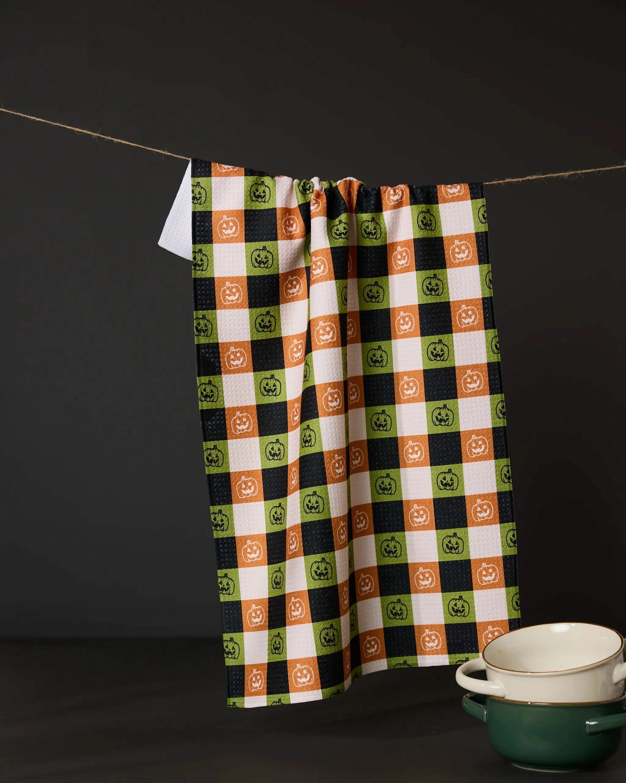 The Jack o Lantern Checker tea towel by Geometry features orange, green, and black checkered squares with pumpkin faces. Made from recycled materials, it hangs above a green pot and white bowls against a dark background.