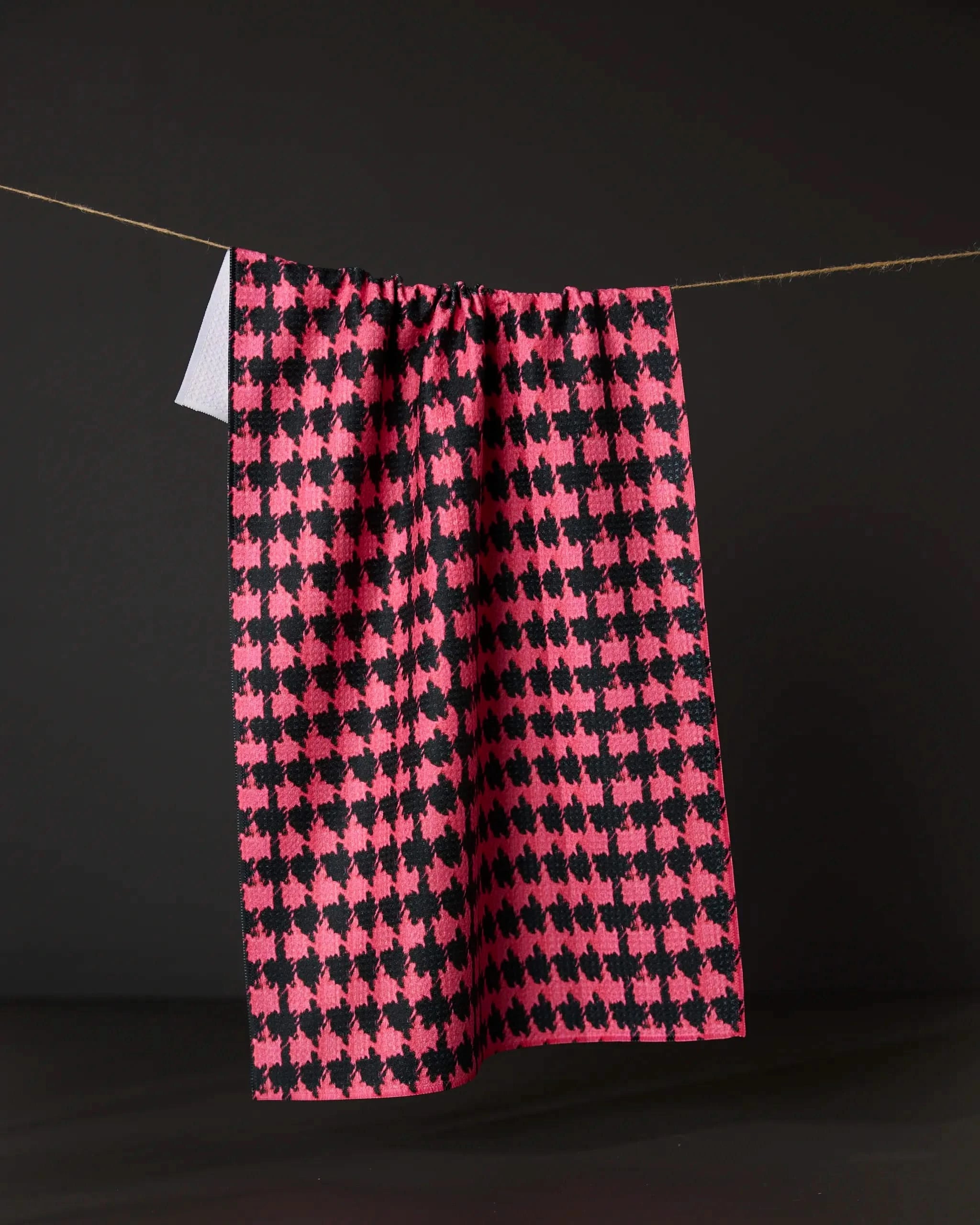 The Geometry Electric Houndstooth dish towel, featuring a pink and black absorbent houndstooth pattern, hangs from a string against a dark background.