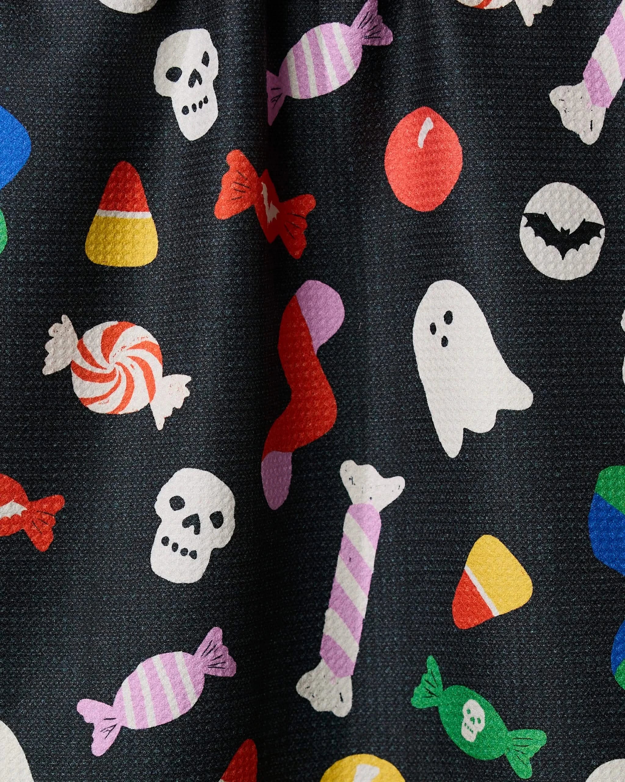 Spooky Candy by Geometry features a festive Halloween pattern of colorful skulls, ghosts, bats, and candy corns on a black fabric. This recycled kitchen towel displays red, yellow, purple, and green sweets in various shapes.