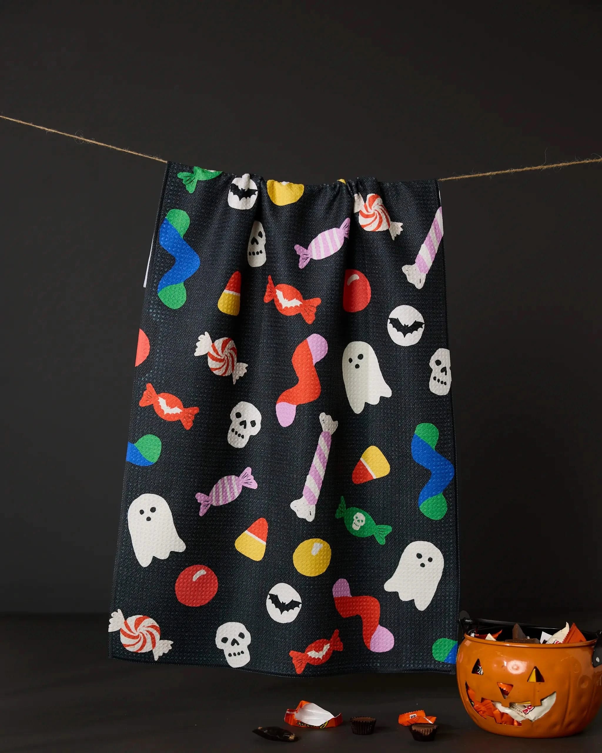 The Geometry Spooky Candy black recycled kitchen towel, featuring vibrant Halloween prints, hangs on a string above a jack-o-lantern bucket filled with assorted candy against a dark background.