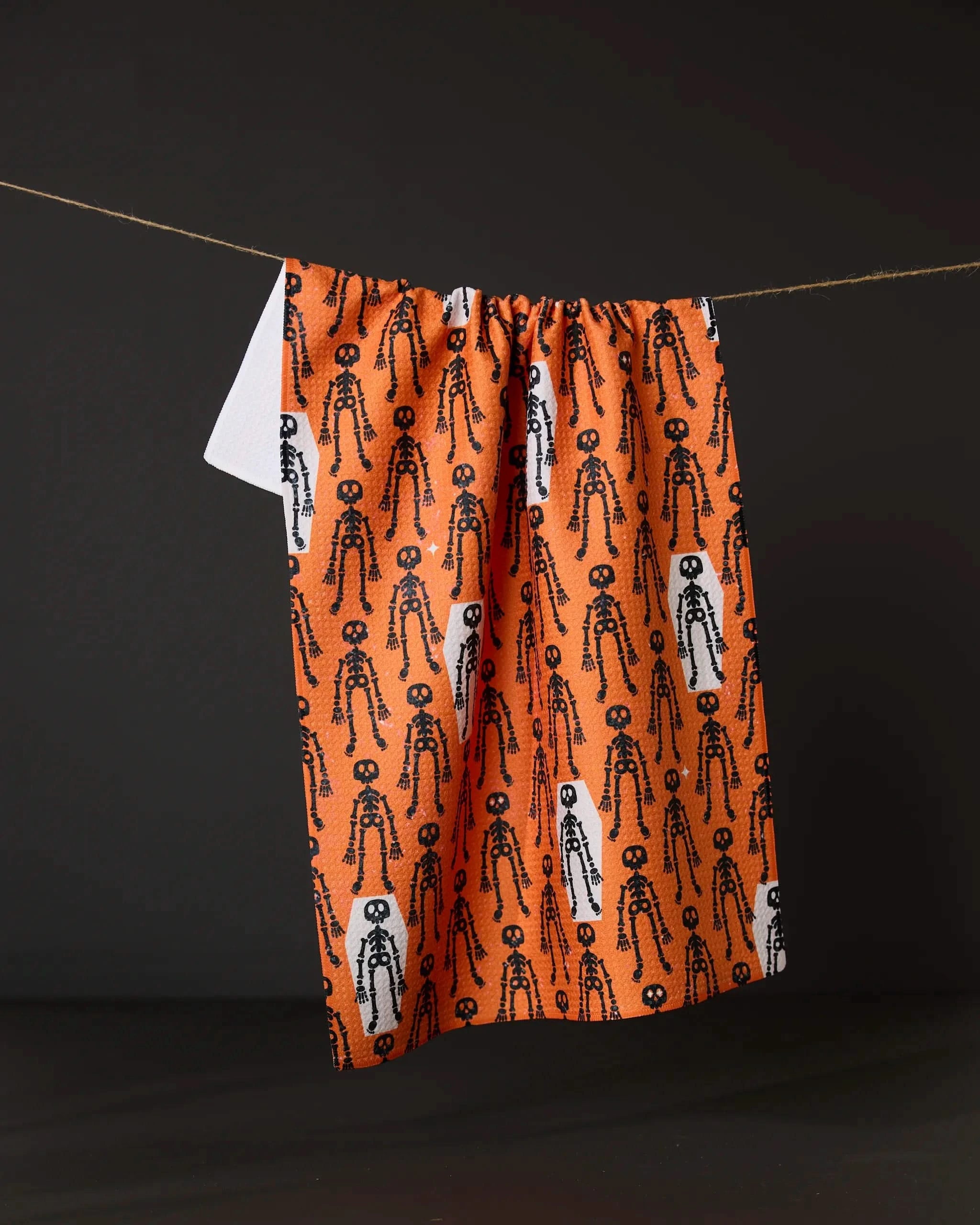 The Resting Skeletons Orange tea towel by Geometry, made from recycled materials, displays a pattern of black and white skeletons and hangs on a string against a dark background.