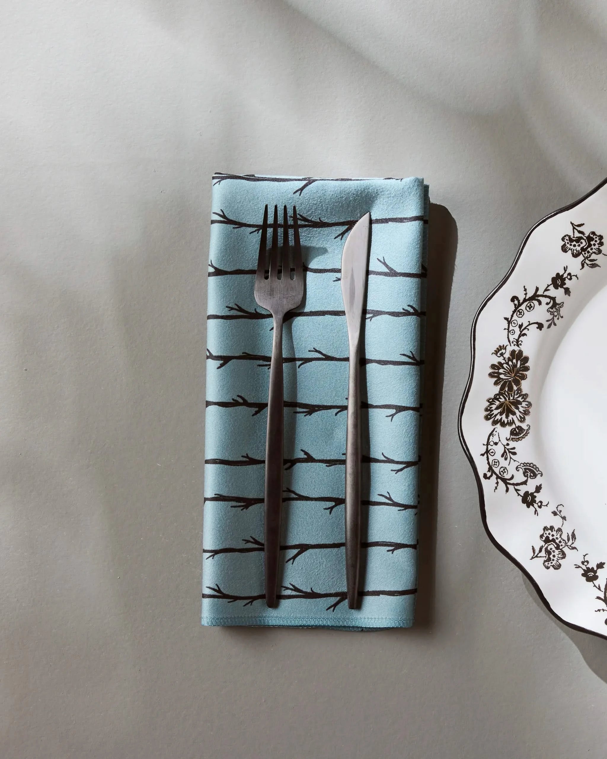 A fork and knife rest on Geometrys Branch Stripes reusable napkin, featuring a black branch pattern, beside a white plate with a black floral design on a light gray surface.