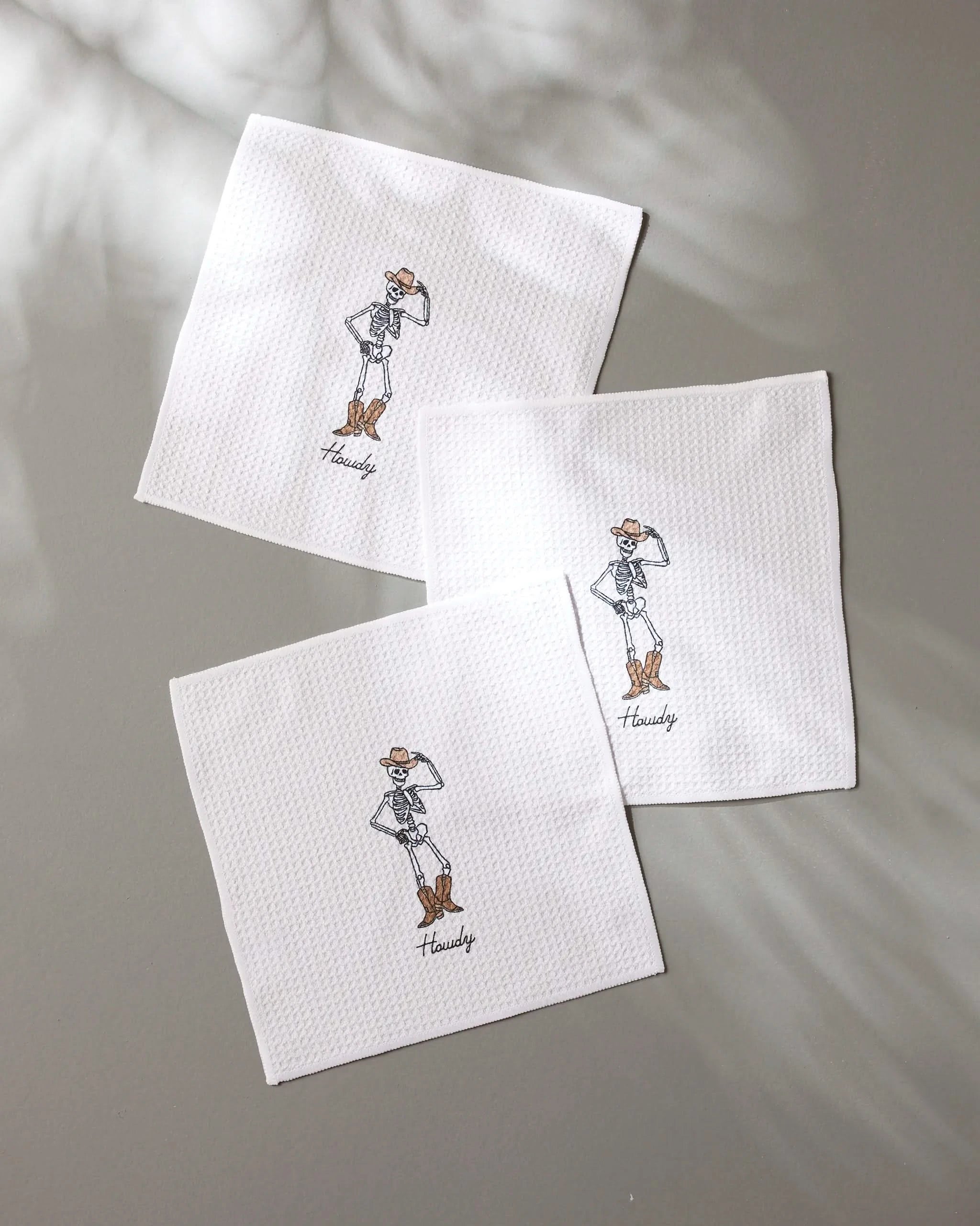 The Geometry Howdy Cassondra set includes three white textured napkins made from recycled materials, each embroidered with a cowboy hat, boots, jacket, and Howdy beneath—combining style with eco-friendly design.