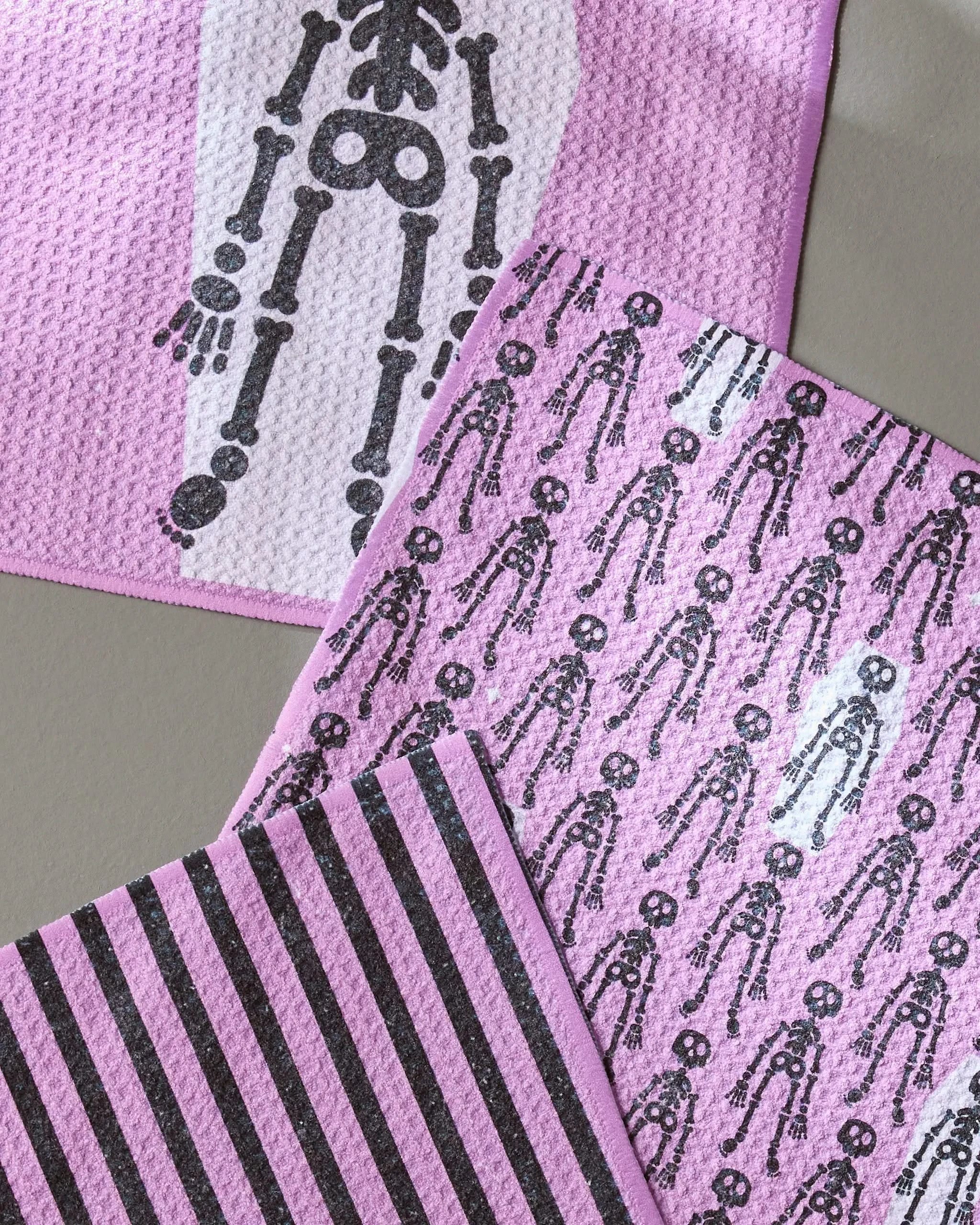 The Geometry Bright Bones kitchen towel set features three pink towels: one with a black skeleton arm, one with a repeating black skeleton print, and one with bold black and purple diagonal stripes.