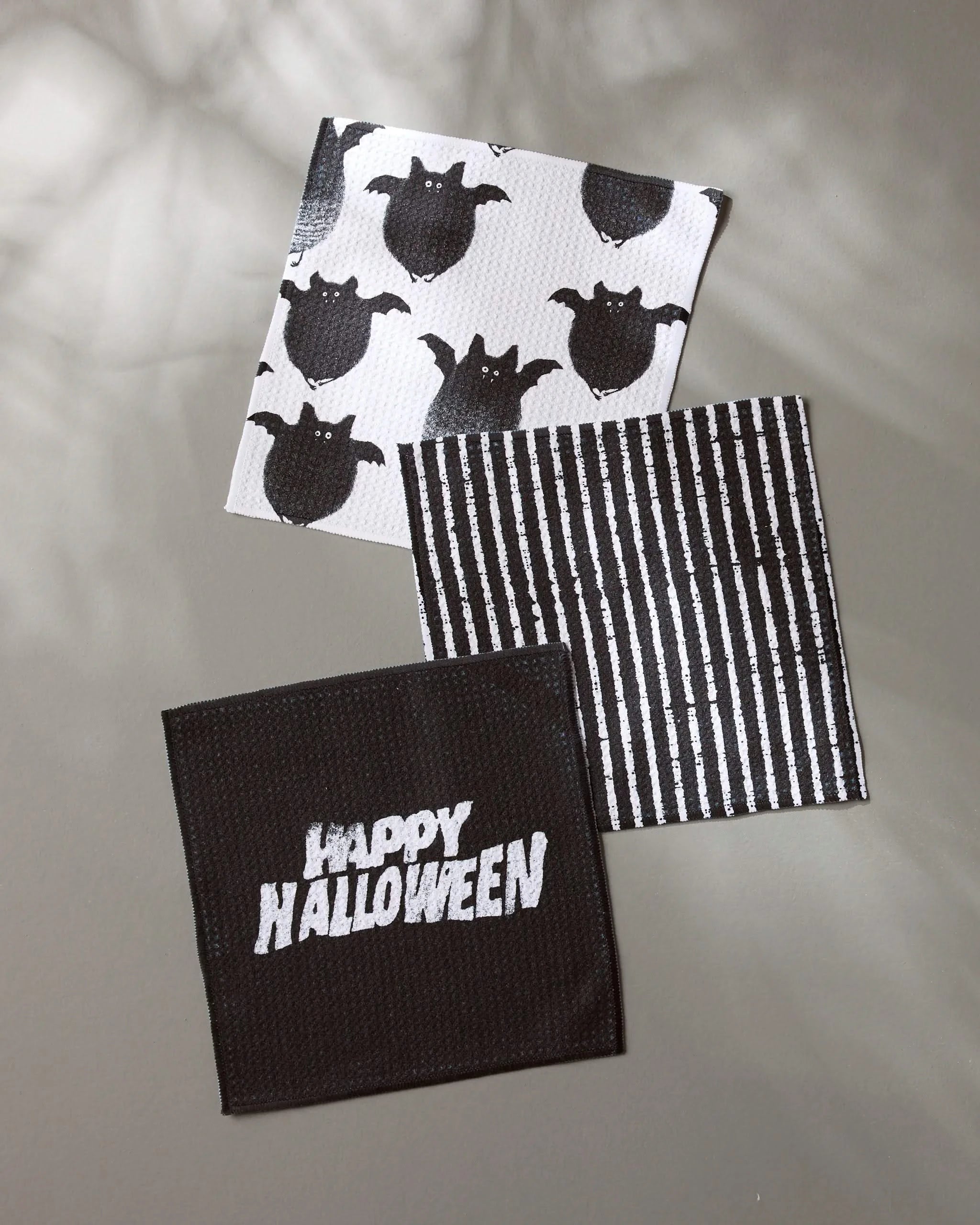 Three square Halloween napkins from Geometry’s Macabre set, made with recycled materials: one with black ghosts, one with black and white stripes, and one that says Happy Halloween in white.