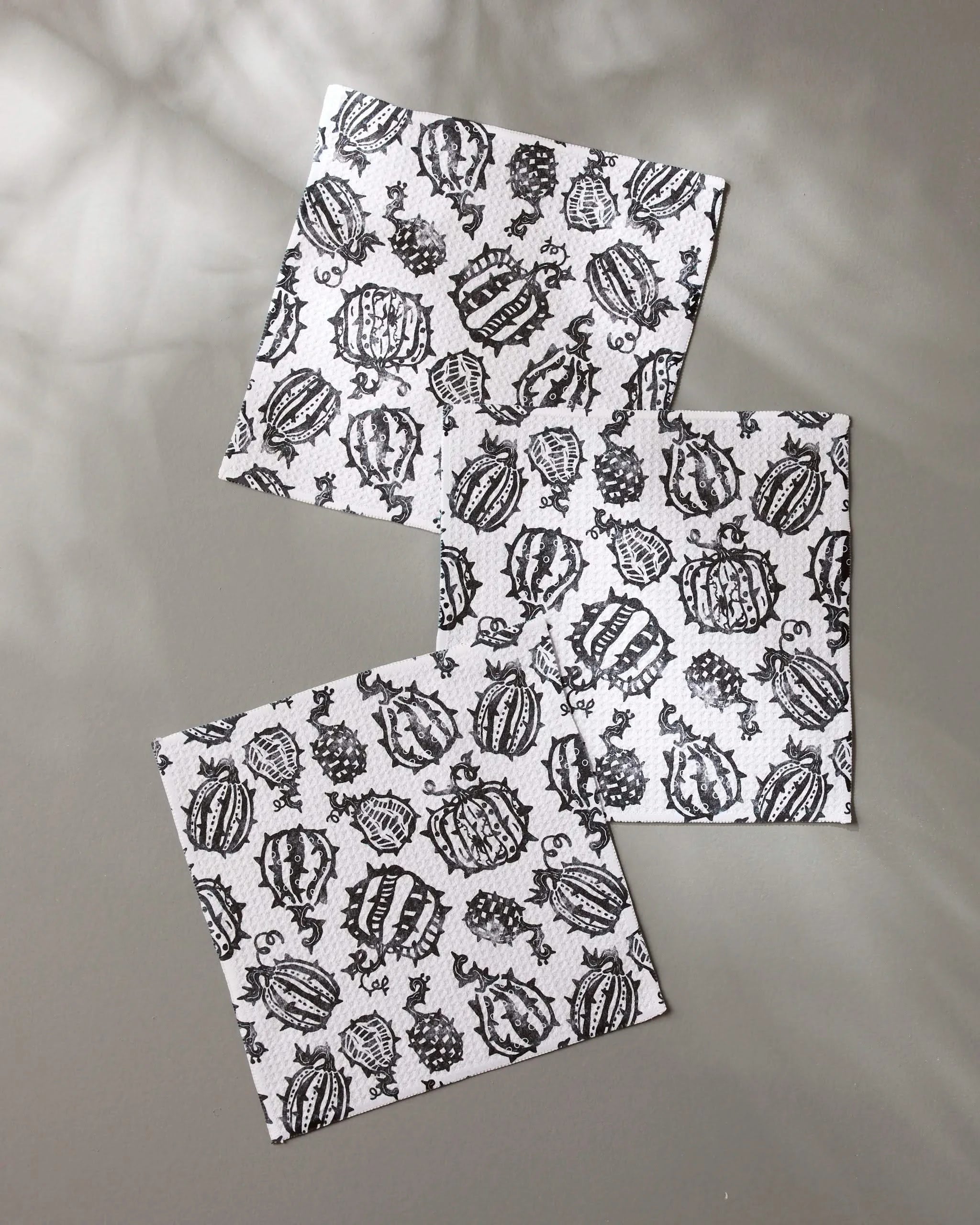 Three Geometry Punk Pumpkins napkins featuring black pumpkin and vine patterns on white, made from recycled materials, are displayed on a gray surface with soft shadows in the background.
