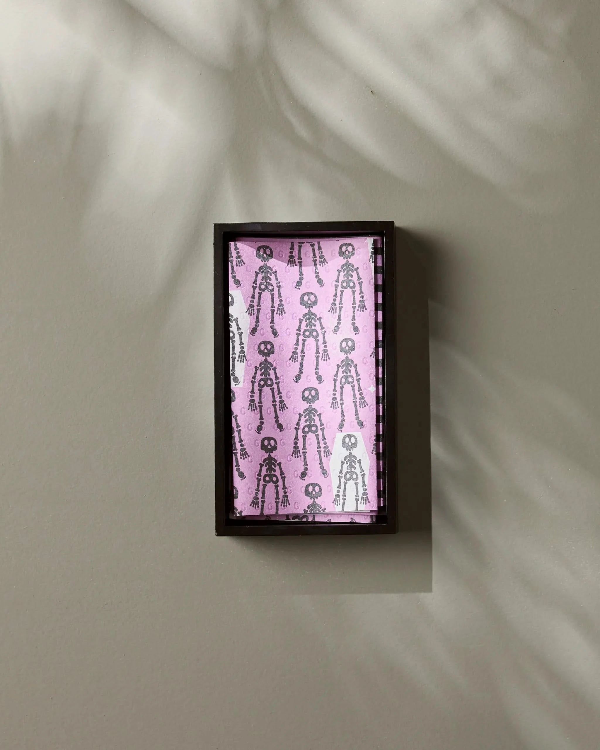 A rectangular black-framed display case by Geometry showcases Bright Bones, featuring a pink background with a repeated black skeleton pattern reminiscent of playful eco-friendly paper towel designs. Soft shadows and light fall across the beige wall.