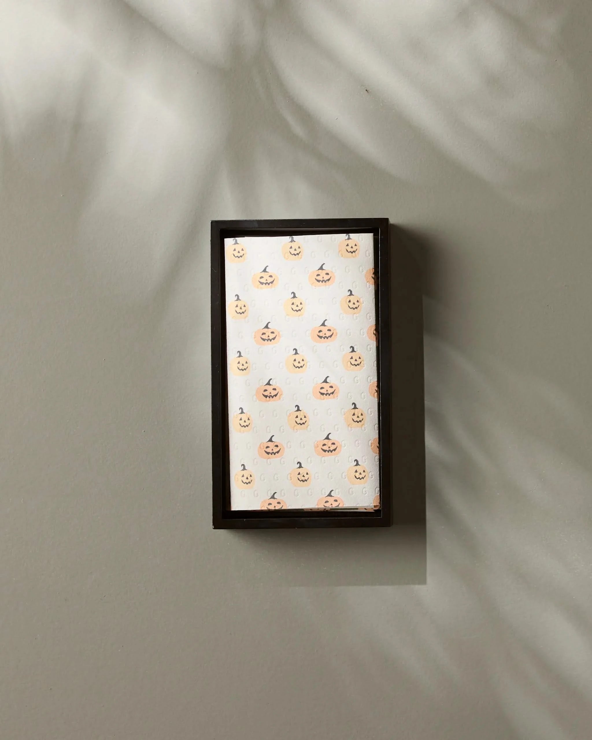 The Spice and Smiles by Geometry is a small, rectangular frame featuring a light orange pumpkin pattern on a white background, inspired by eco-friendly kitchen towels. It hangs on a neutral wall with leafy shadows softly cast across it.