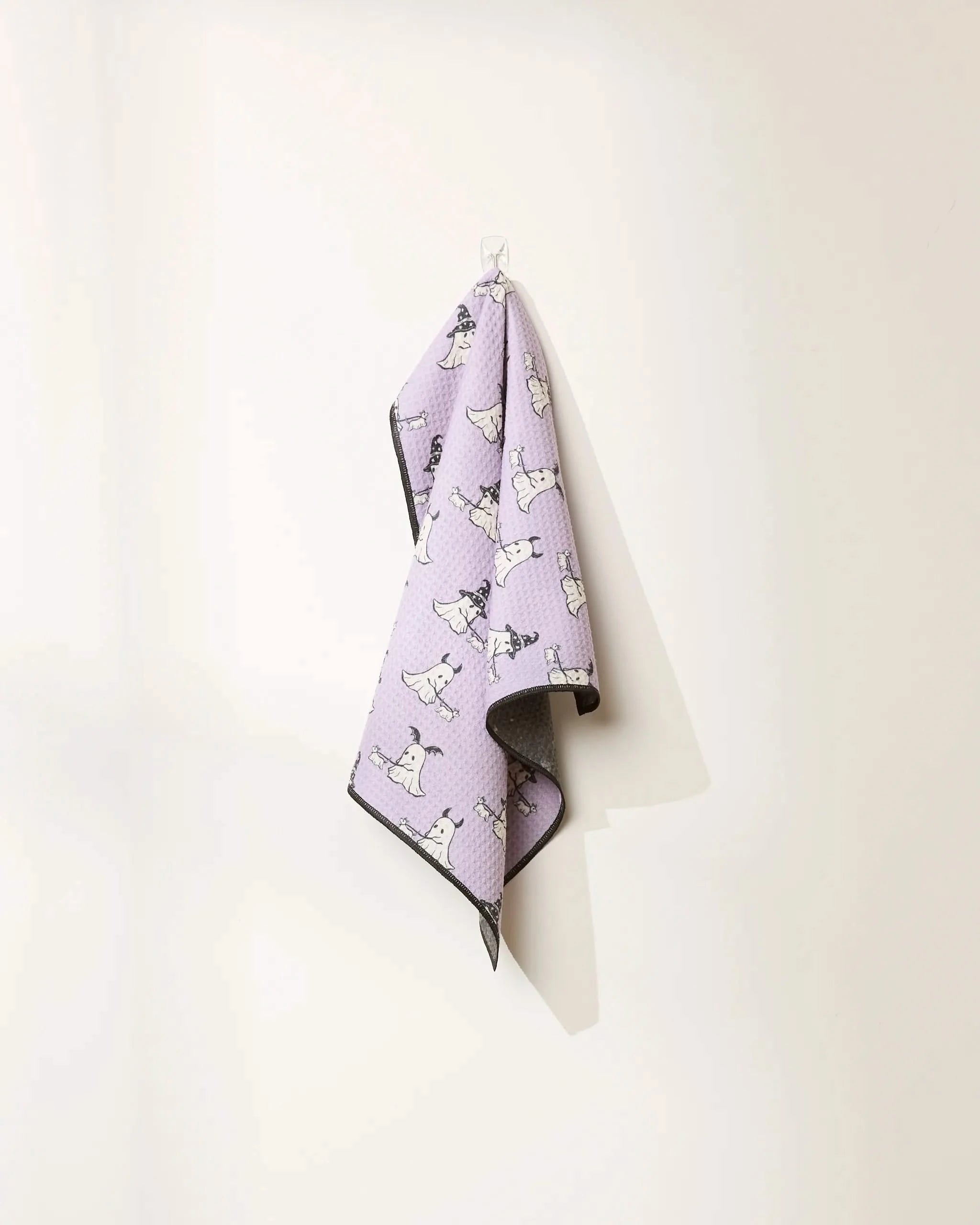 A Spooky Ghost Walk Paw Towel by Geometry with a cartoon ghost design hangs from a clear hook on a light wall. The ultra-absorbent towel has dark edges and casts a shadow.