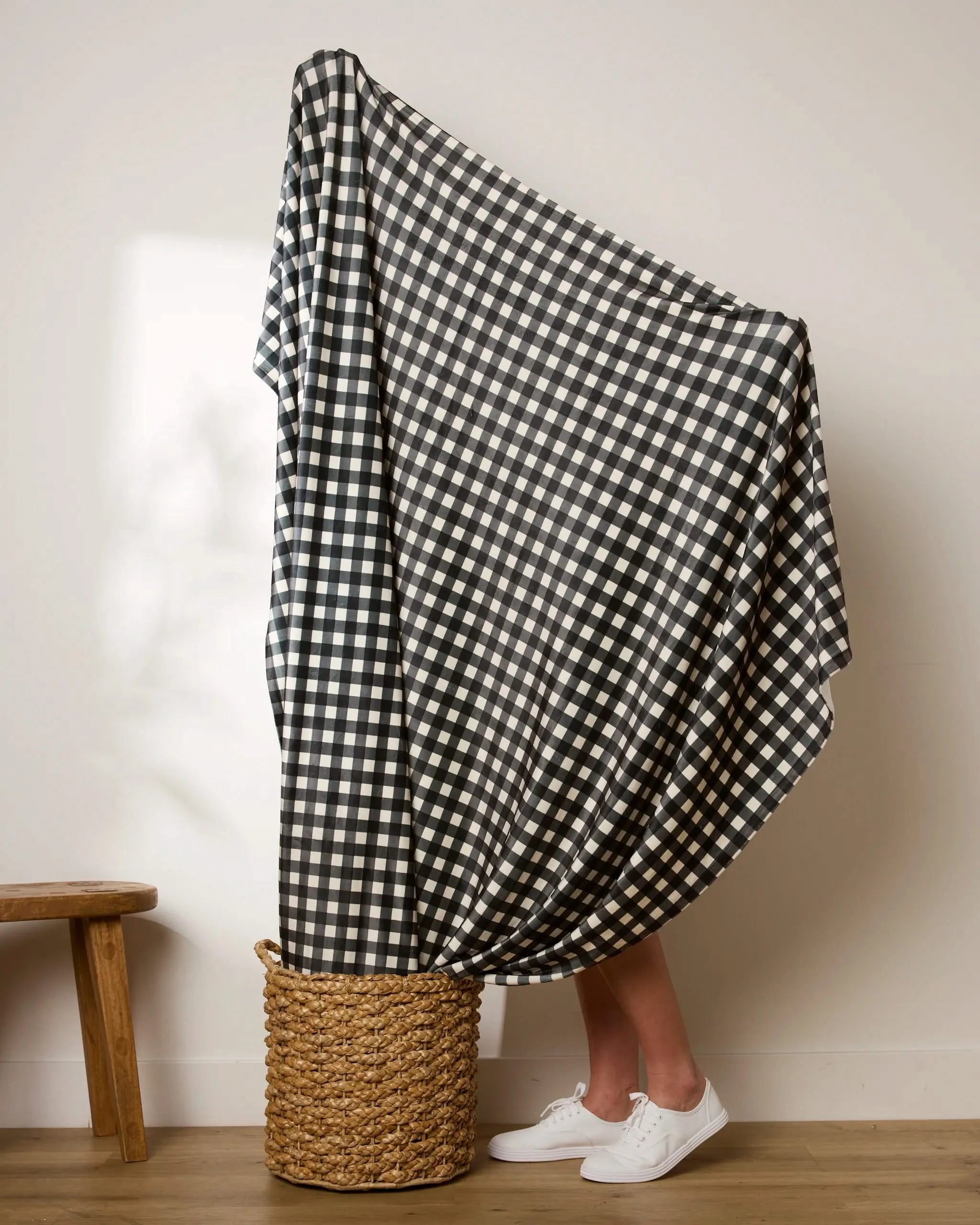 A person stands in a basket draped with the Geometry Boohoo Charcoal blanket, its bold black-and-white checkered pattern contrasting against their white sneakers; a small wooden bench sits nearby by the plain wall.