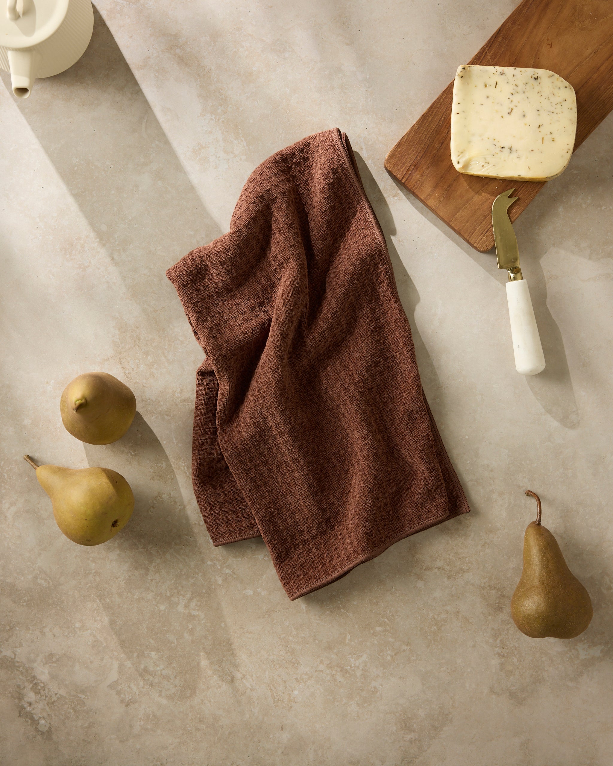 The Geometry Maple kitchen towel in brown texture is displayed on a stone countertop with three yellow pears, a wedge of herb cheese on a wooden board, a cheese knife, and part of a white teapot.