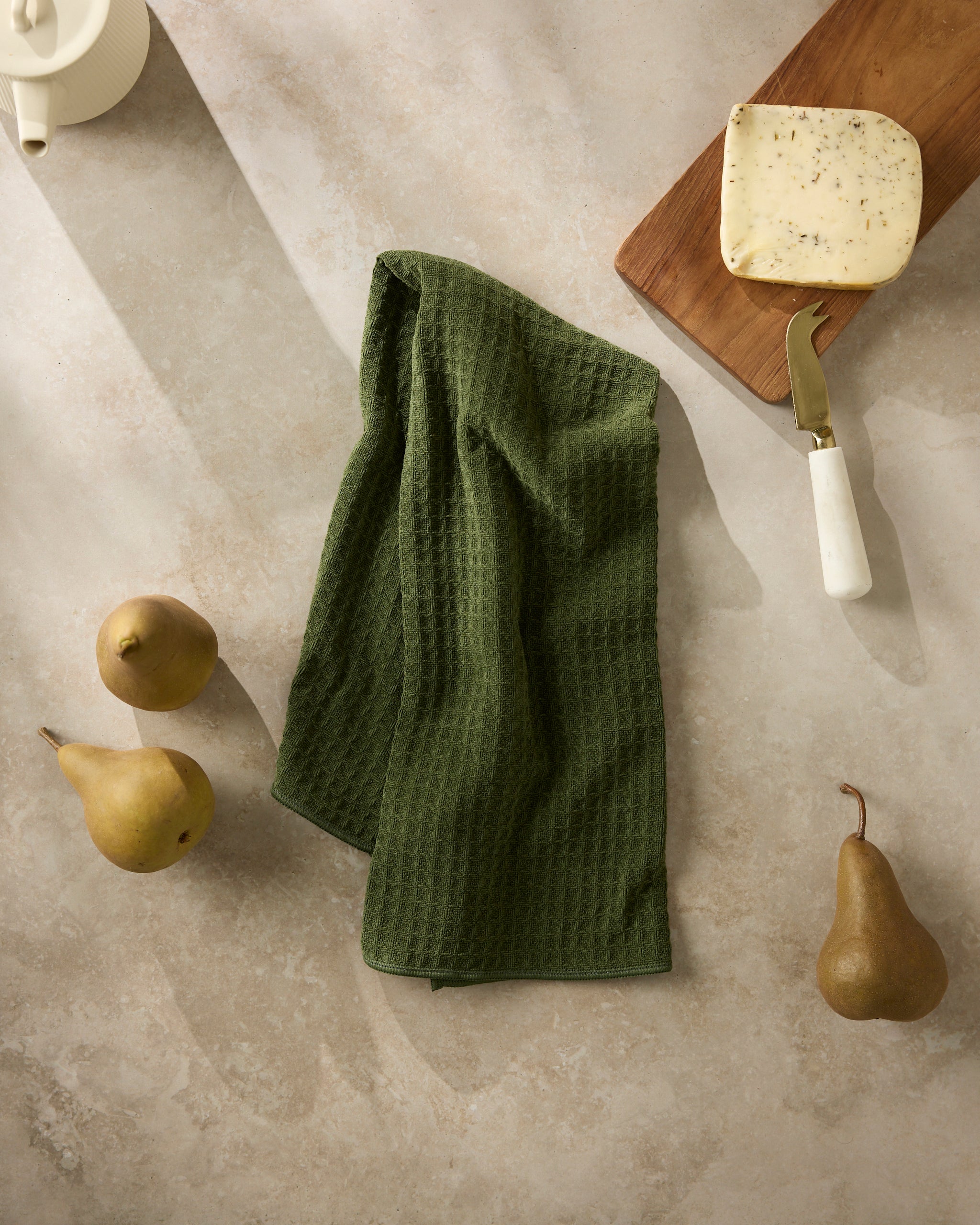 A Geometry Matcha textured kitchen towel is laid on a light stone countertop next to three pears, herb cheese on a wooden board, and a white-handled cheese knife. Sunlight casts gentle shadows across the scene.