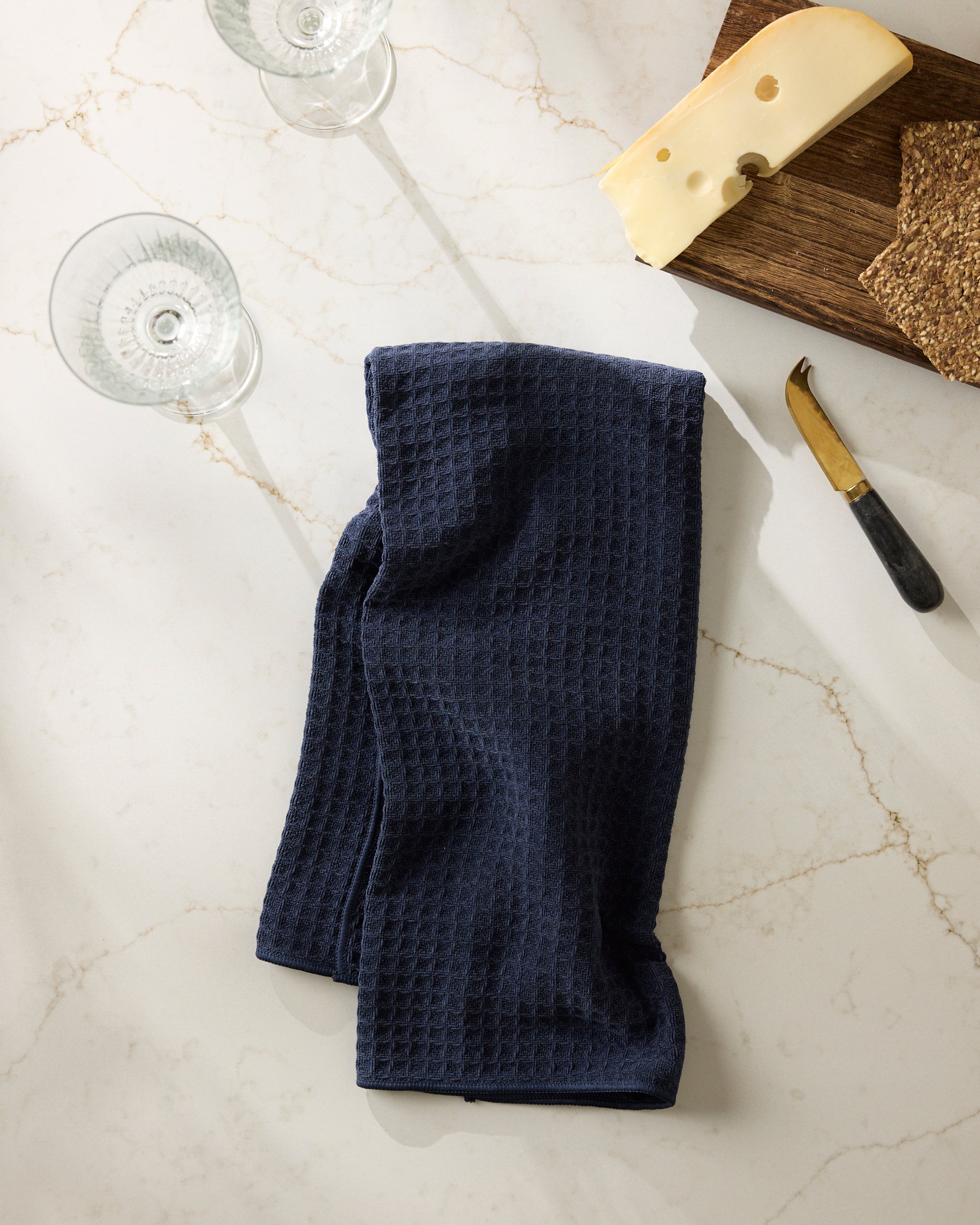 The Geometry Sapphire dark blue textured kitchen towel rests on a white marble countertop beside two empty wine glasses, a block of Swiss cheese on a wooden board, a cheese knife, and some crackers.