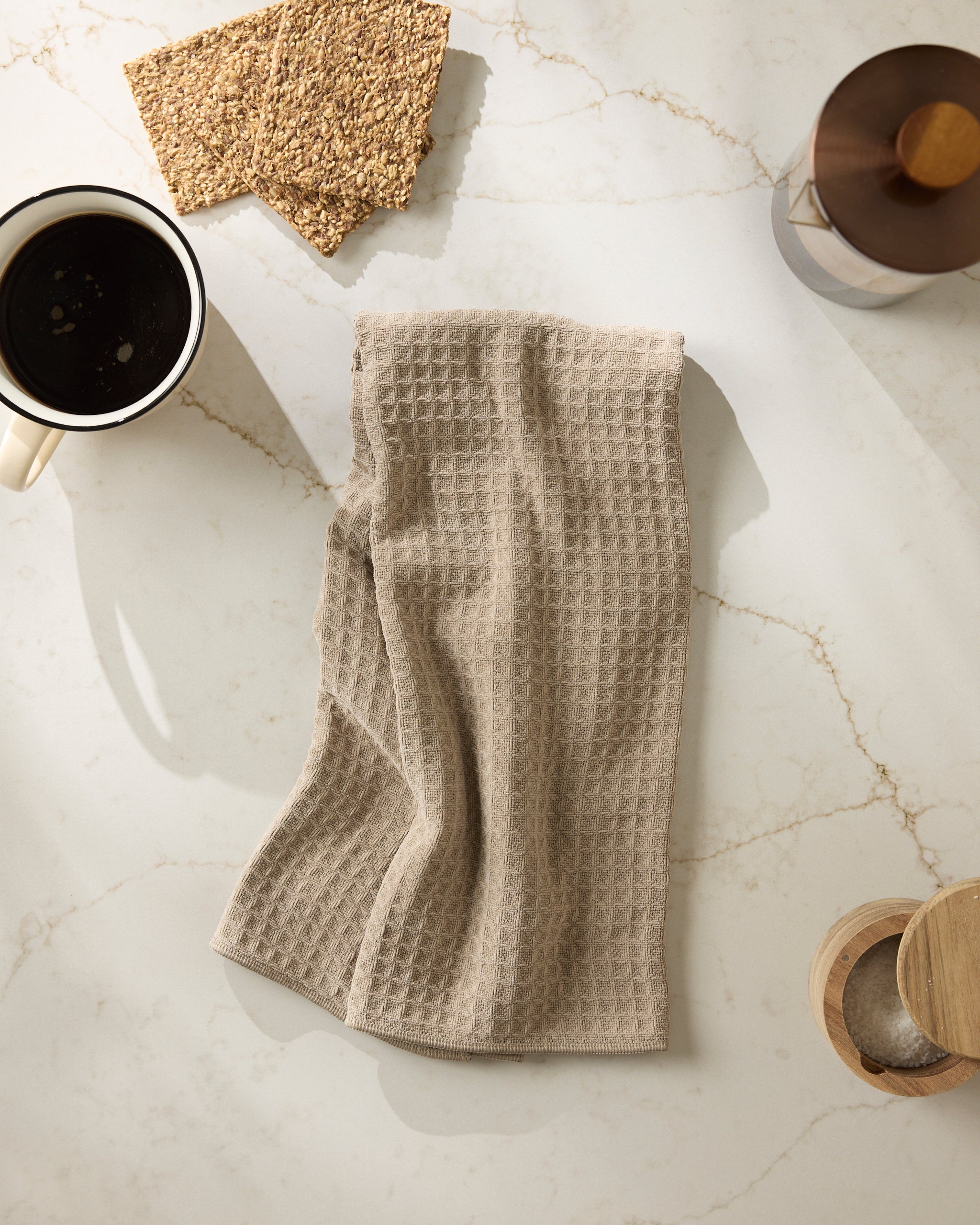 The Geometry "Latte" kitchen towel with a beige waffle texture is displayed on white marble near a cup of black coffee, crispbread, and wooden utensils in soft natural light.