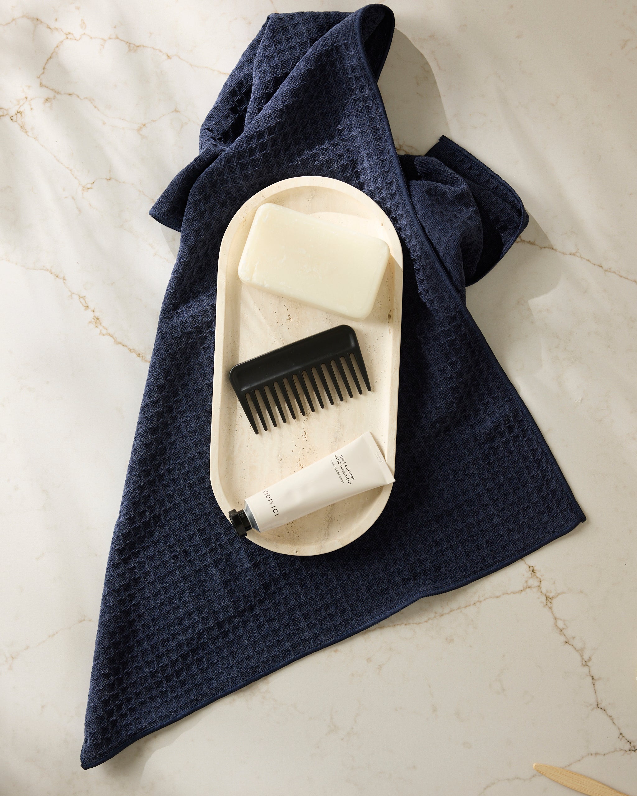 The Geometry "Sapphire" towel in navy blue, displayed with a wooden tray holding a soap bar, black comb, and white cream tube, set on a light marble surface.