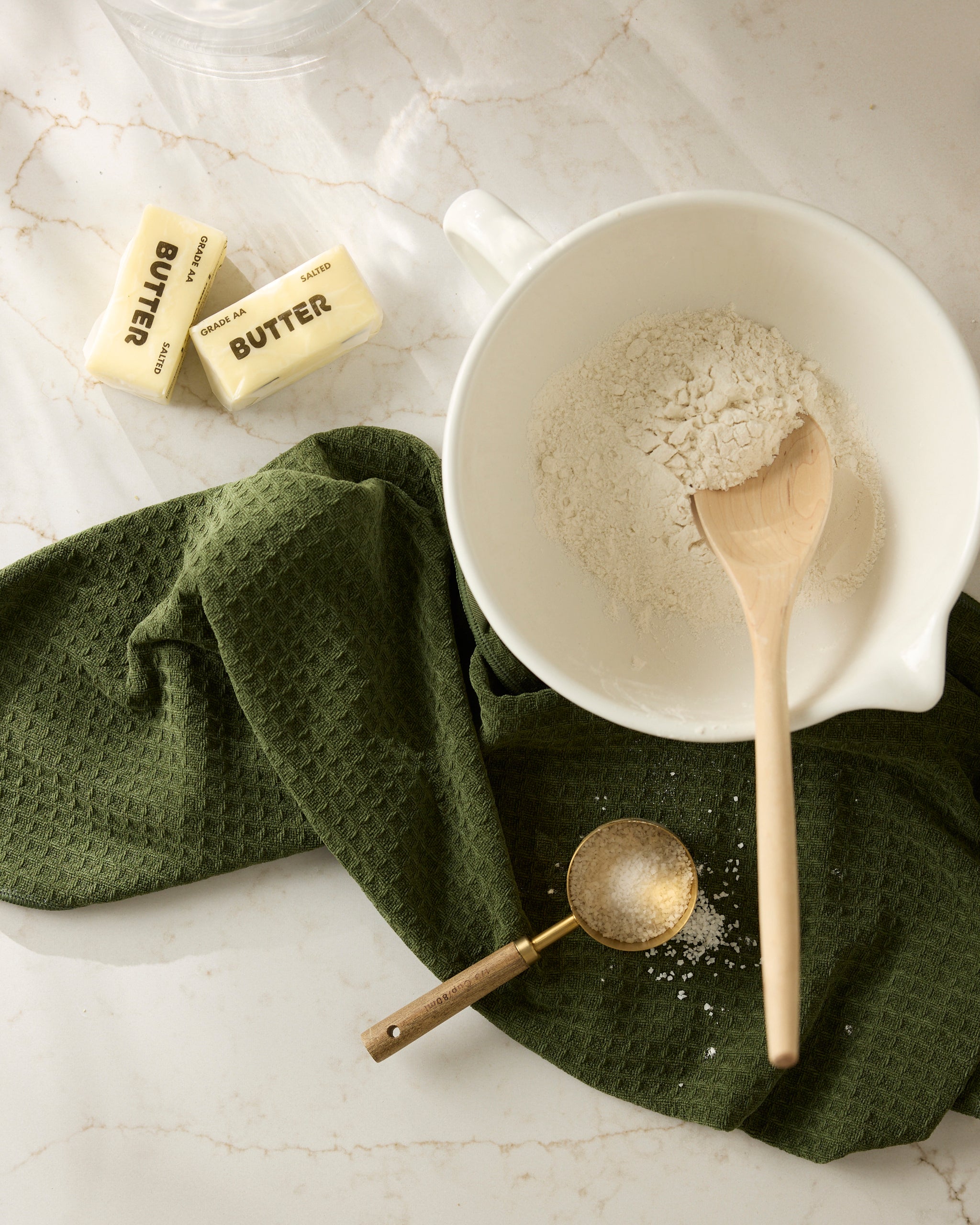 A bowl with Matcha flour and a wooden spoon sits on a green Geometry cloth. Nearby are two sticks of butter and a metal measuring spoon with flour, all on a light marble countertop.