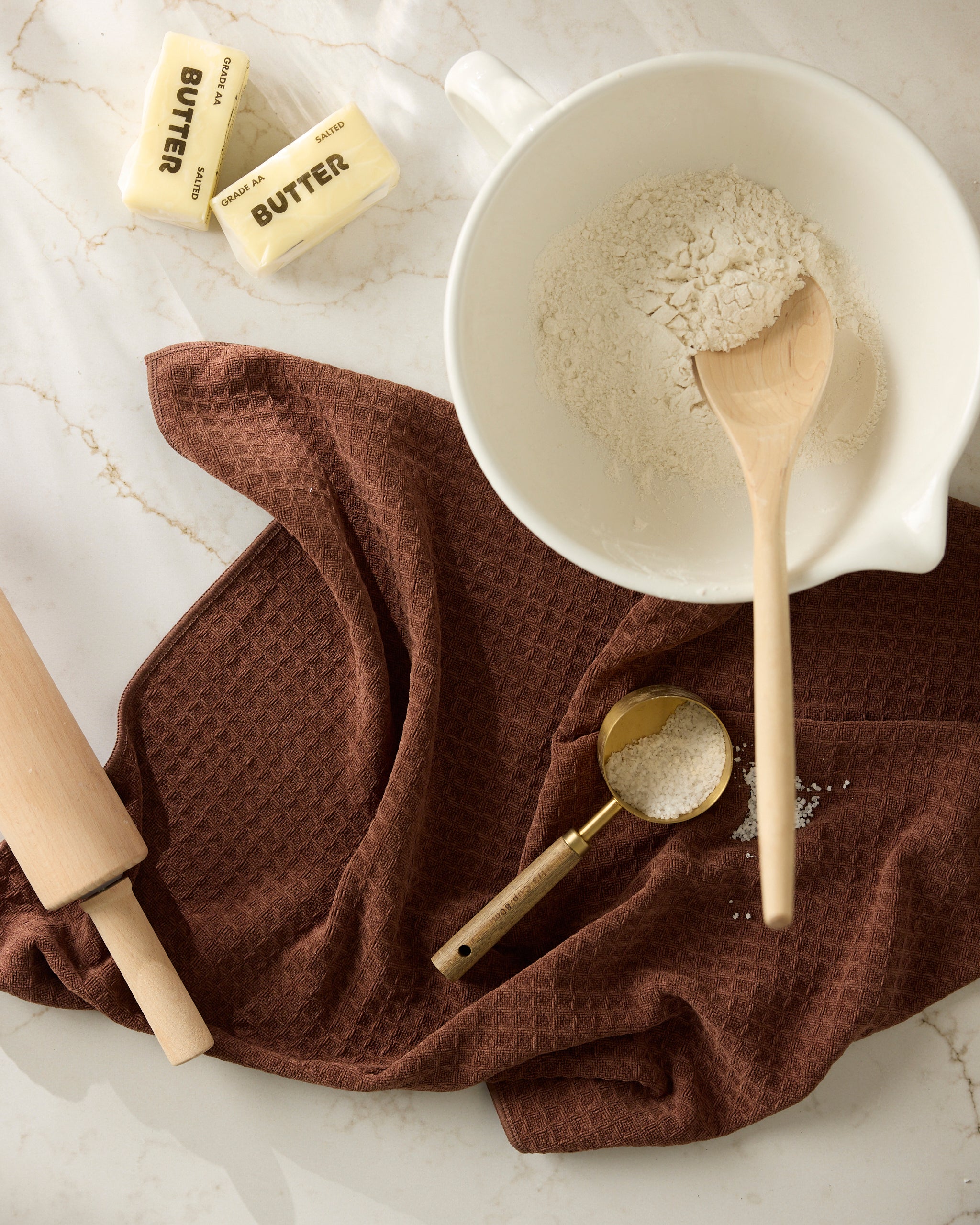 The Geometry Maple mixing bowl with flour and a wooden spoon rests on a brown kitchen towel, alongside two butter sticks, a rolling pin, and a gold measuring cup of flour on the marble countertop.