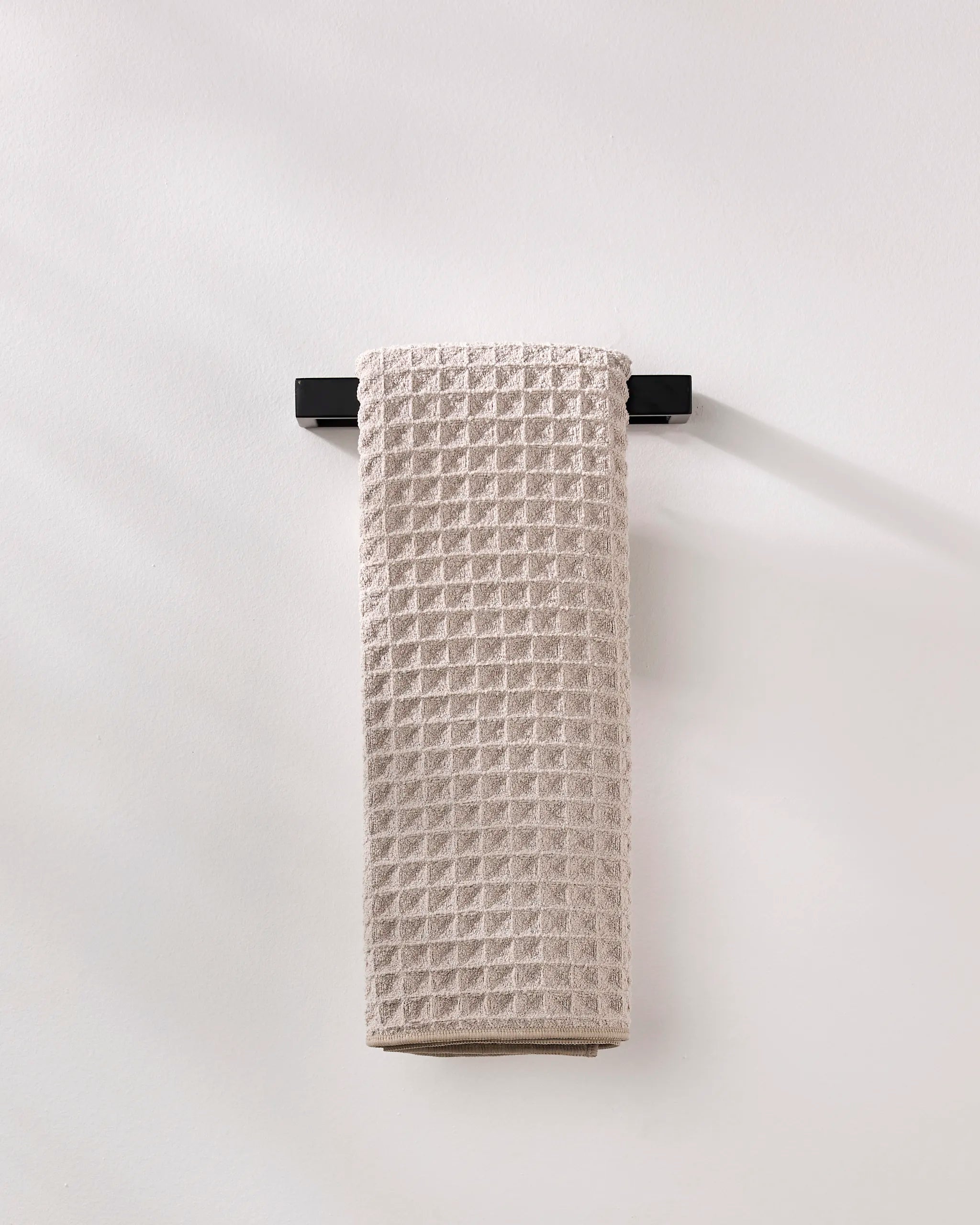 A GEOMETRY Taupe waffle hand towel made from recycled materials hangs neatly on a sleek black towel bar against a plain white wall.