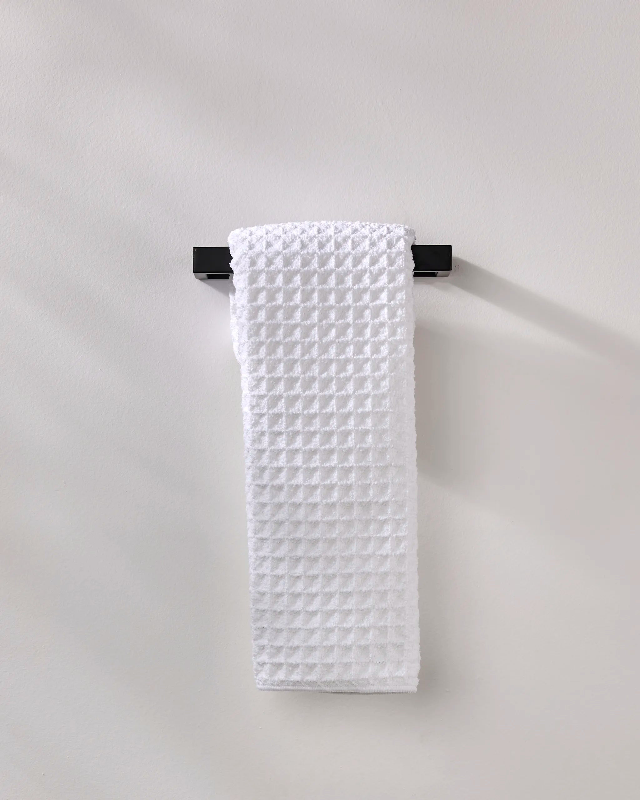 The GEOMETRY Cloud hand towel, made from recycled materials, in white waffle texture, hangs neatly on a black towel bar fixed to a light gray wall.