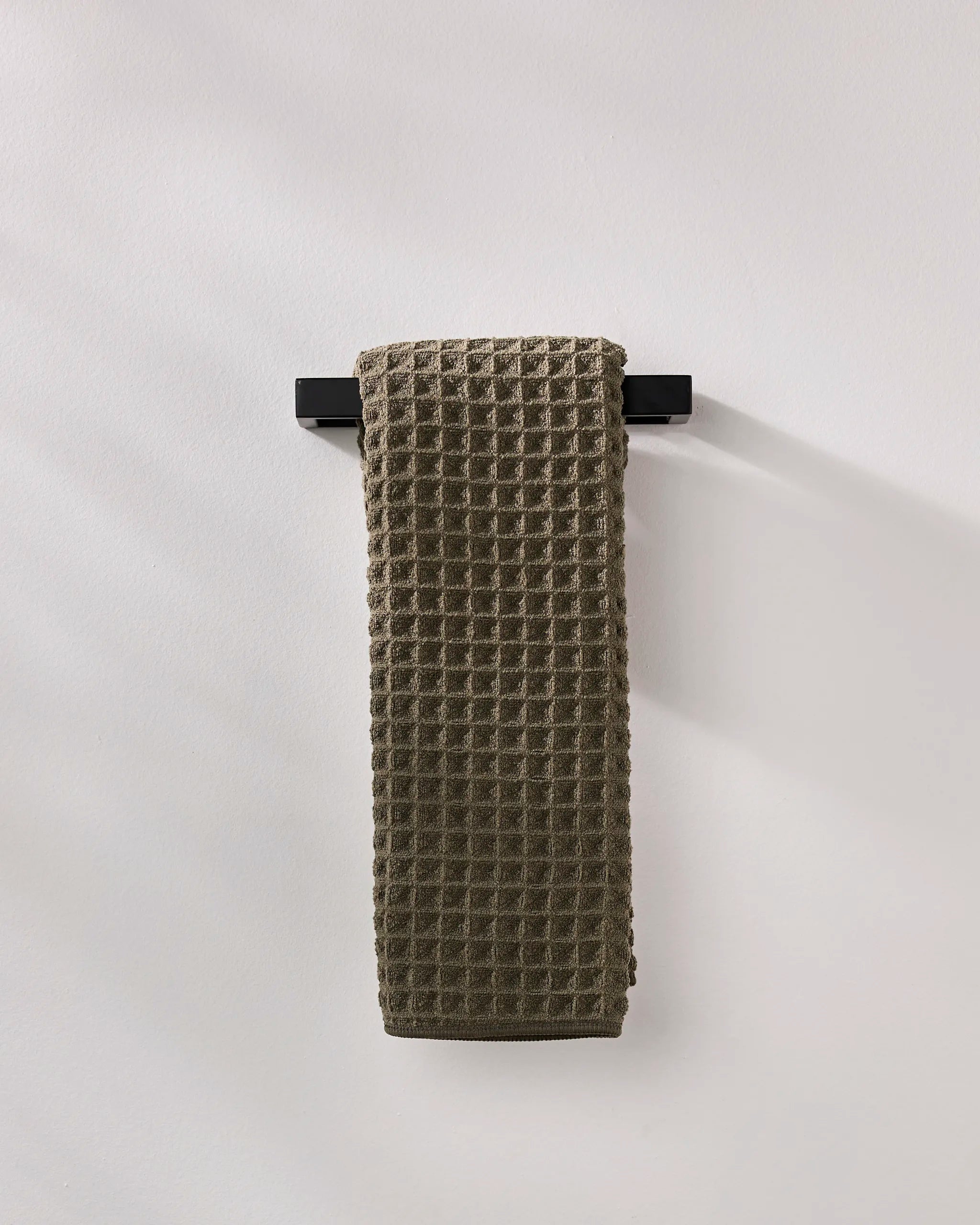 A GEOMETRY Forest ultra-absorbent waffle hand towel in dark gray hangs neatly on a black towel bar against a plain, light-colored wall.