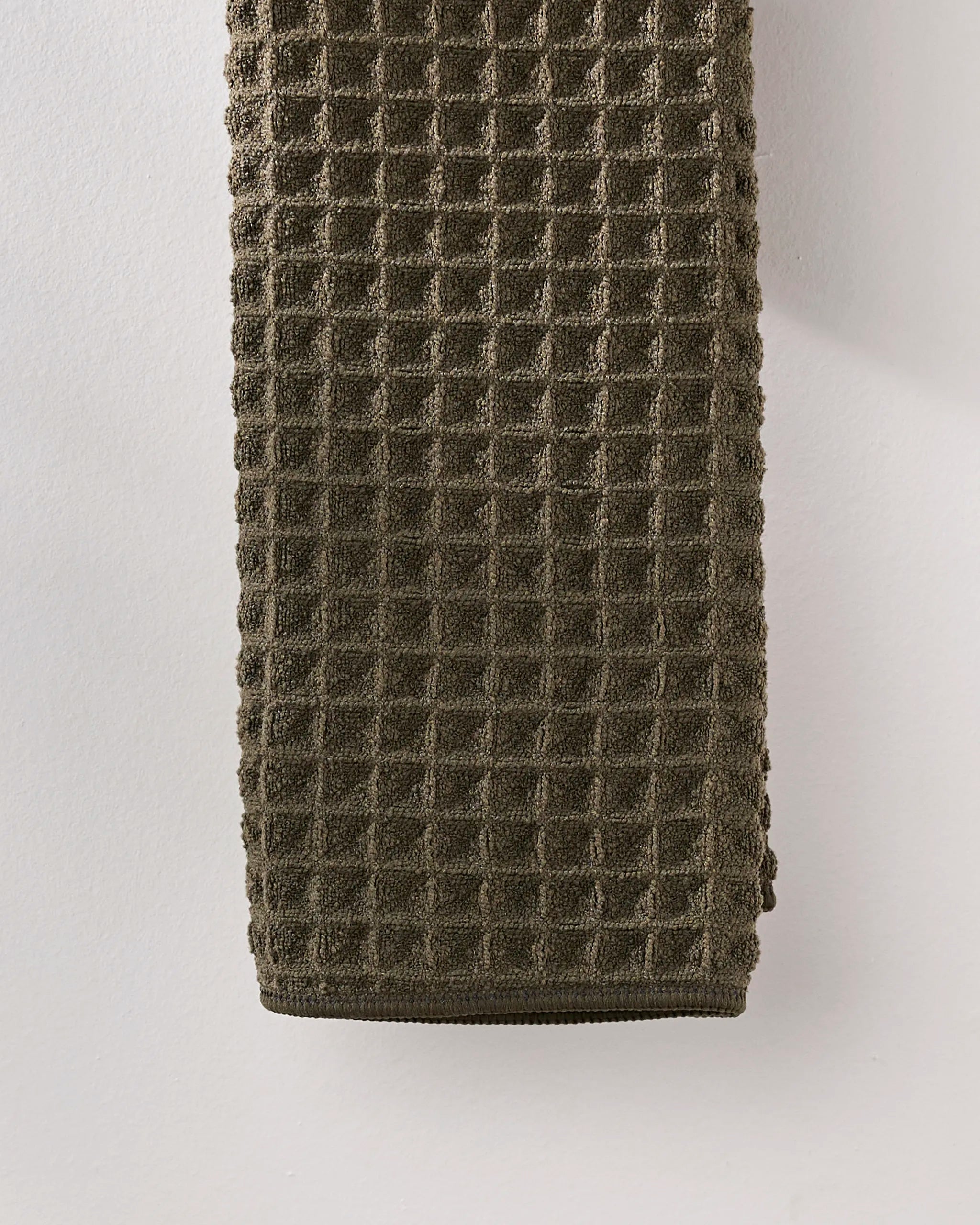 The GEOMETRY "Forest" hand towel in dark olive green features a textured waffle weave and hangs against a light wall, adding an ultra-absorbent touch to your bath space.