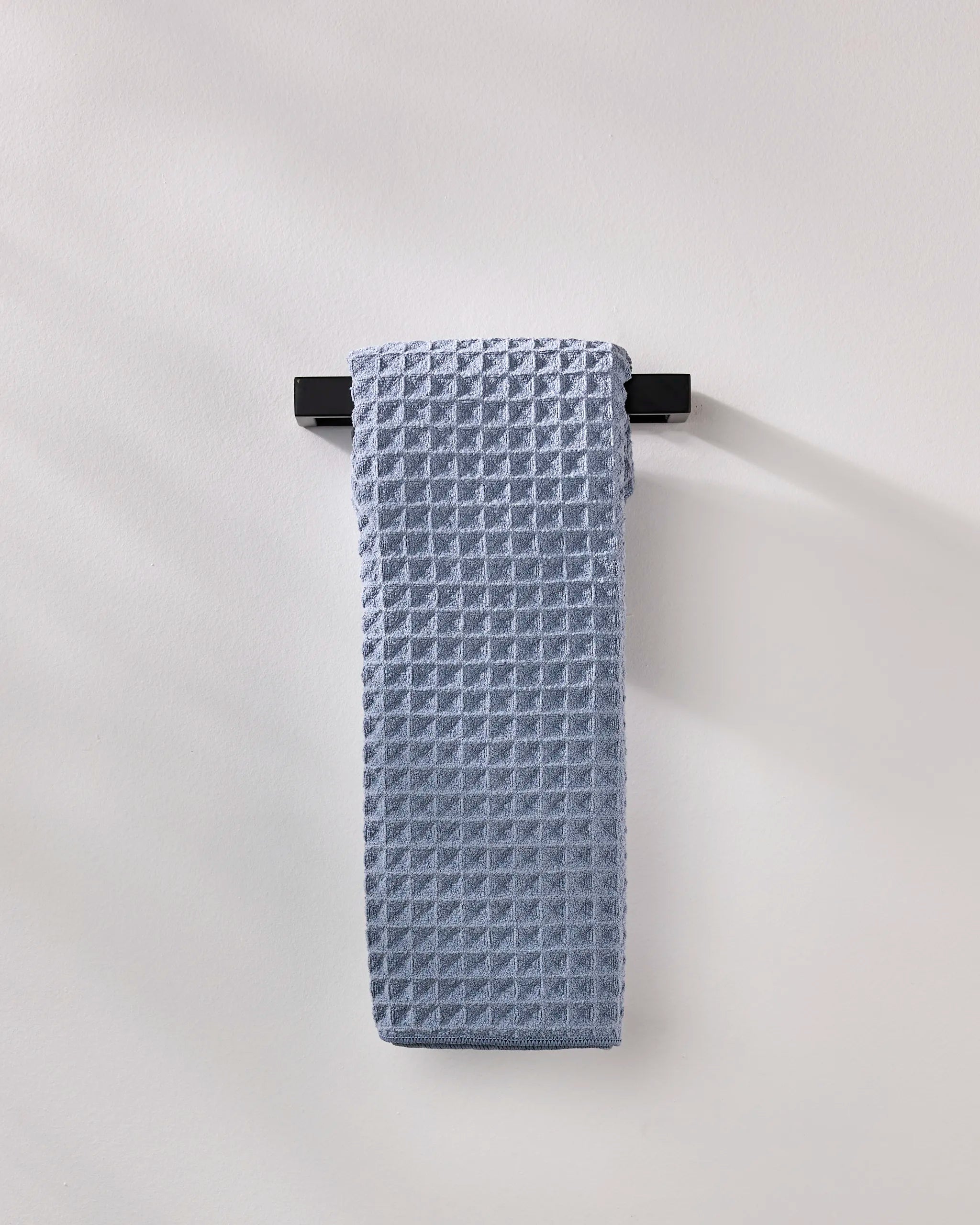 The GEOMETRY Sky hand towel, neatly folded in light blue waffle texture, hangs on a sleek black bar against a plain wall for an understated style touch.
