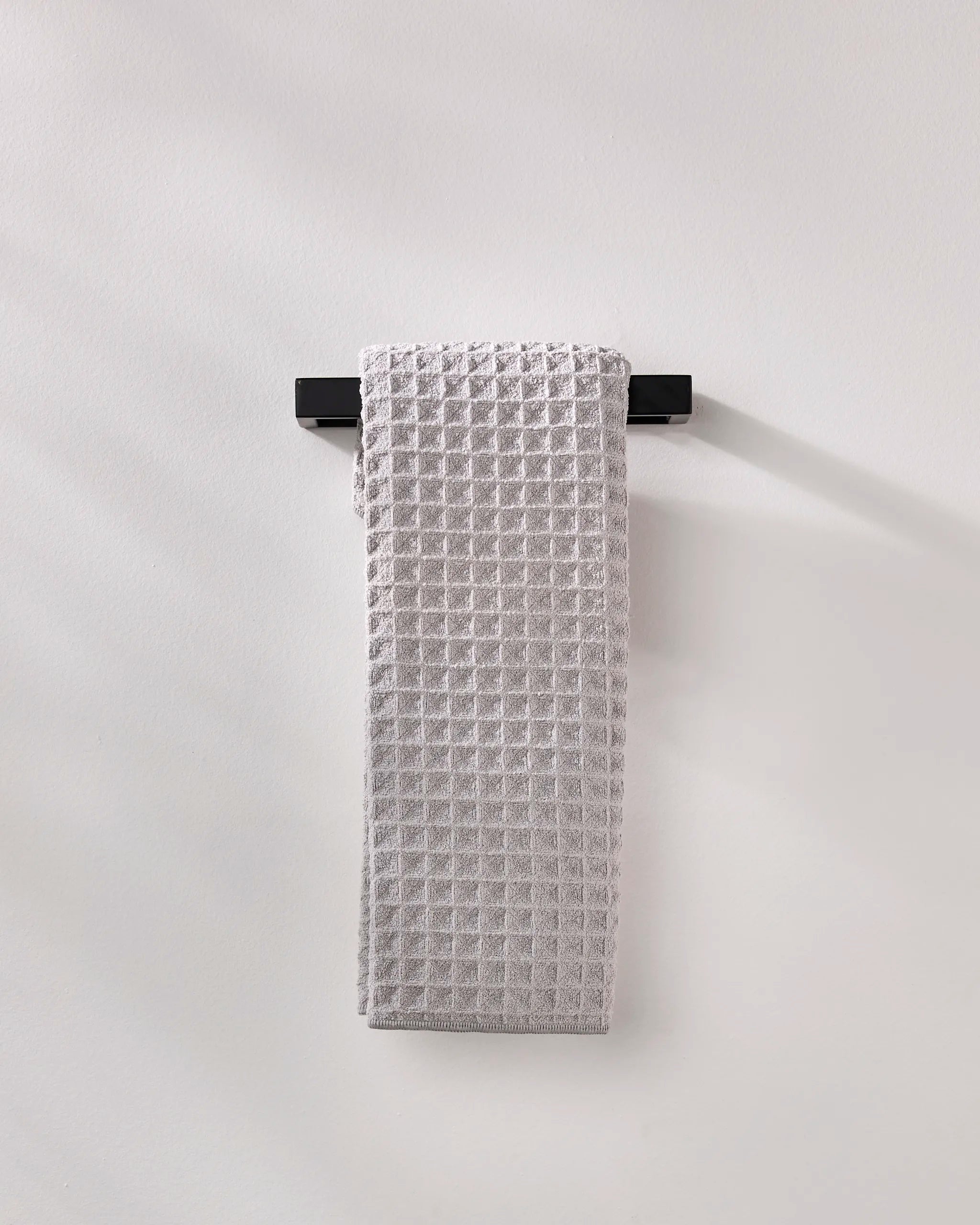 A light gray, waffle-textured GEOMETRY "Stone" luxury bath towel is neatly draped over a sleek black towel rack mounted on a plain, light-colored wall.