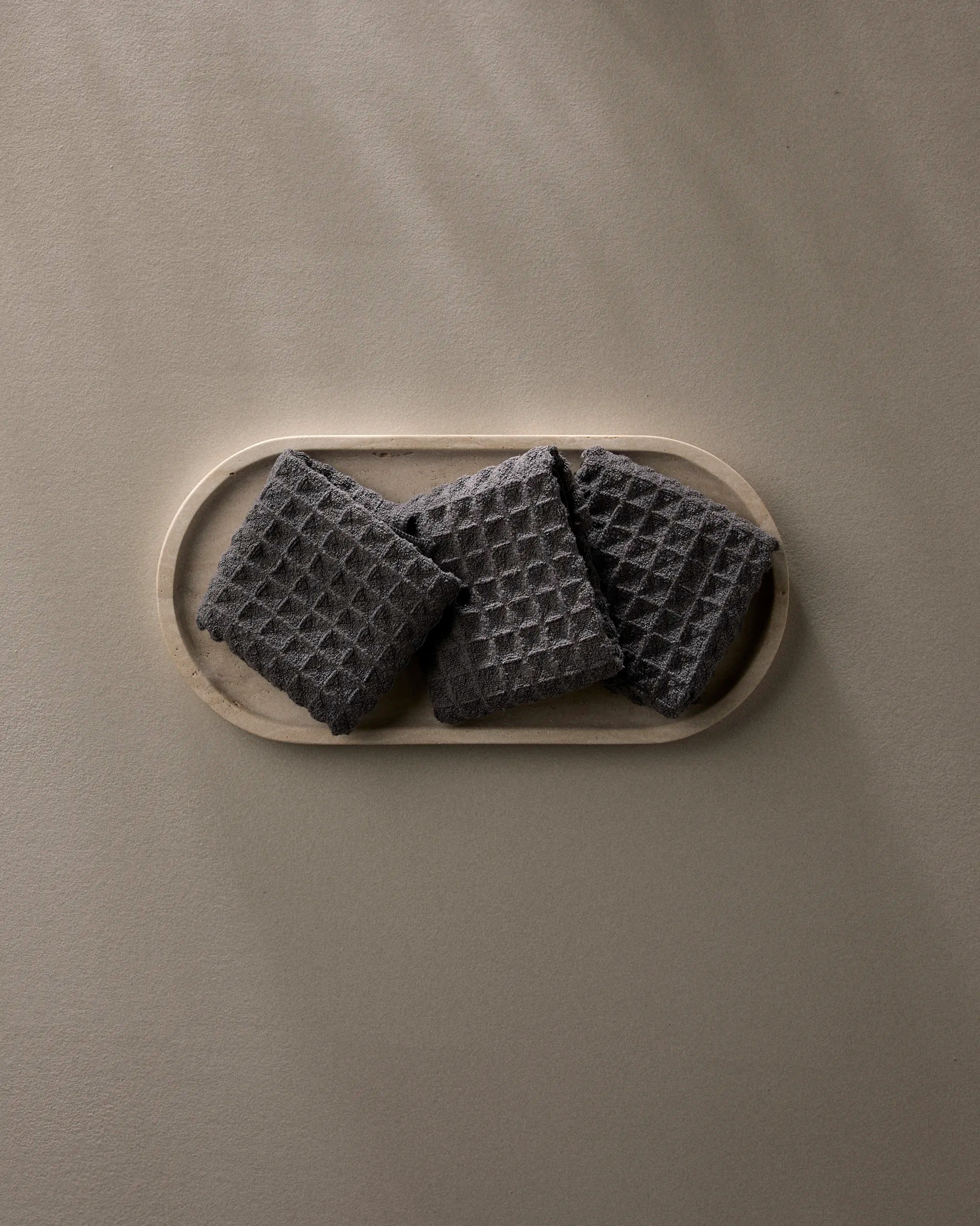A wooden tray displays three GEOMETRY Charcoal waffle washcloths, neatly arranged on a light beige surface, with soft natural light creating gentle shadows.