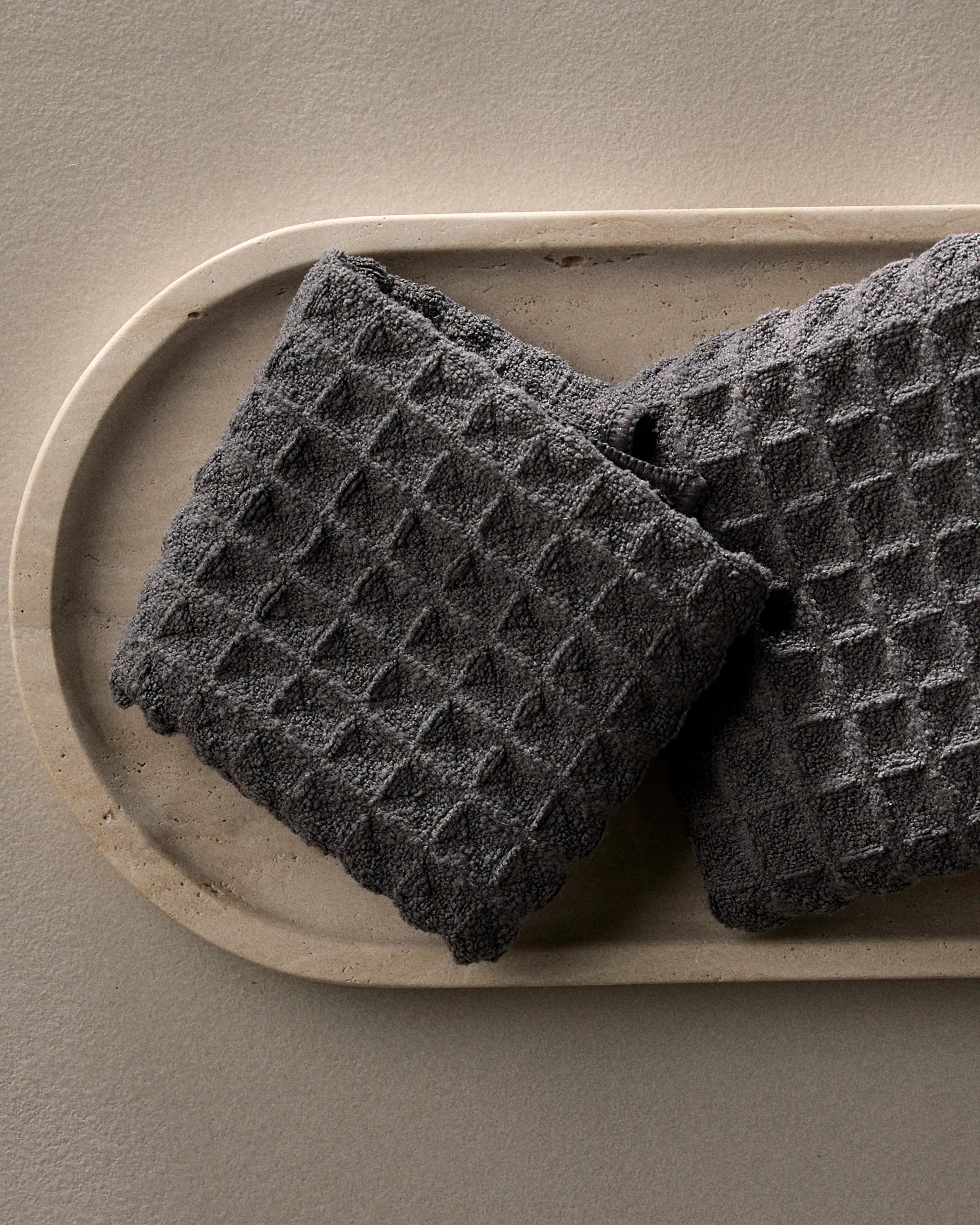 Two GEOMETRY “Charcoal” waffle washcloths with a diamond pattern are neatly placed on a beige, oval tray atop a light taupe surface.
