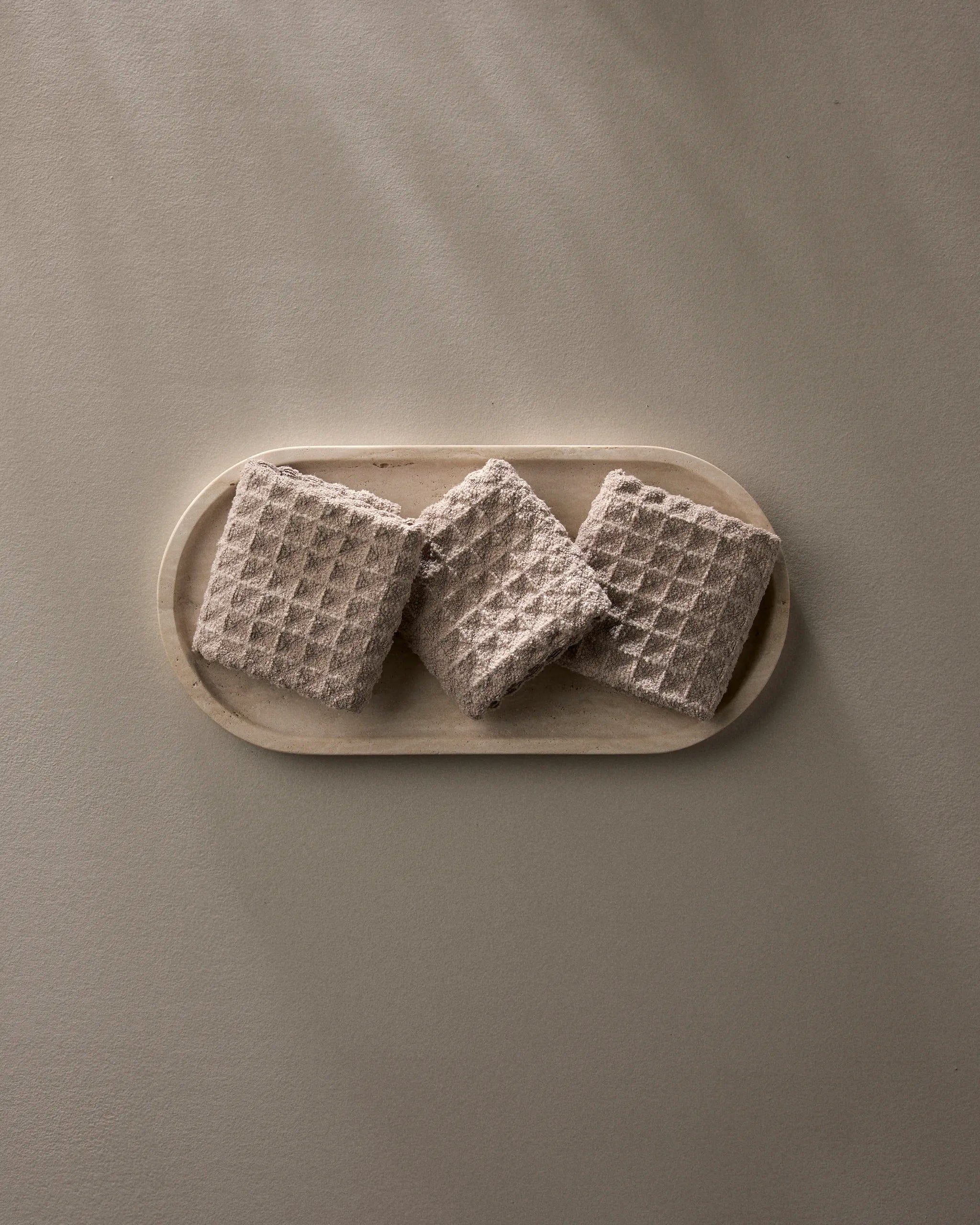 A Taupe oval plate by GEOMETRY displays three square, textured crackers with a waffle pattern—arranged side by side—on a neutral surface.