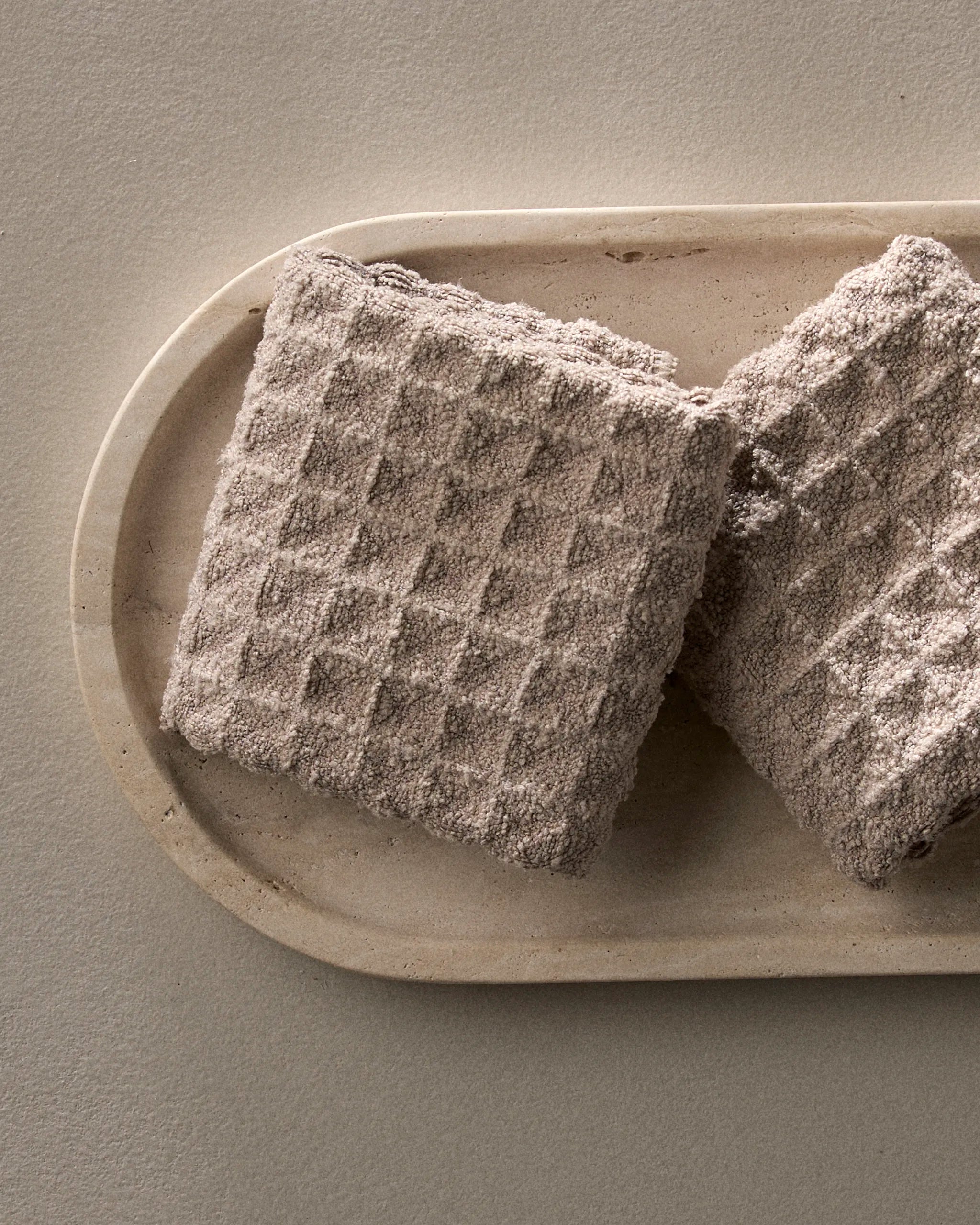 Two square, textured Taupe wafers by GEOMETRY with a dusty light brown coating rest on an oval beige ceramic plate against a neutral background.