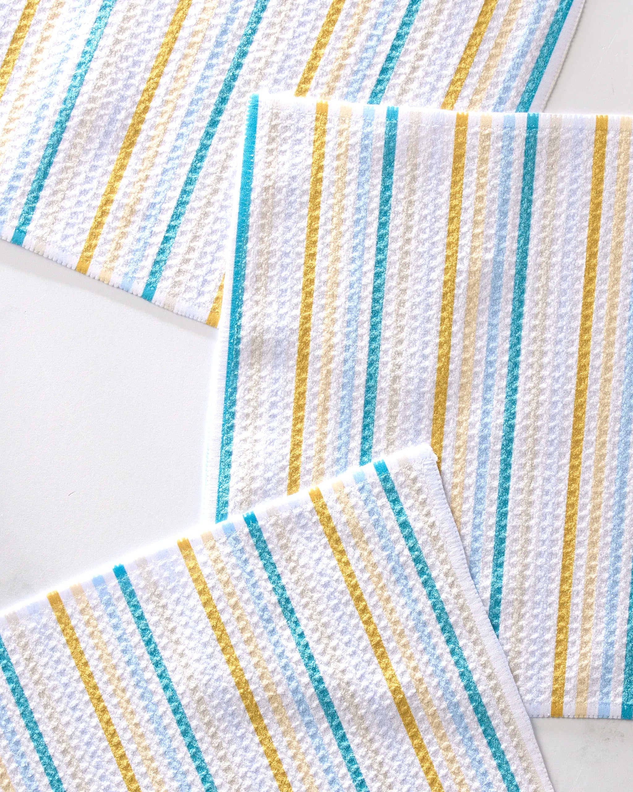 Three GEOMETRY Seaside Sunshine white textured recycled dishcloths with yellow, teal, and gray diagonal stripes are partially overlapping on a white surface.