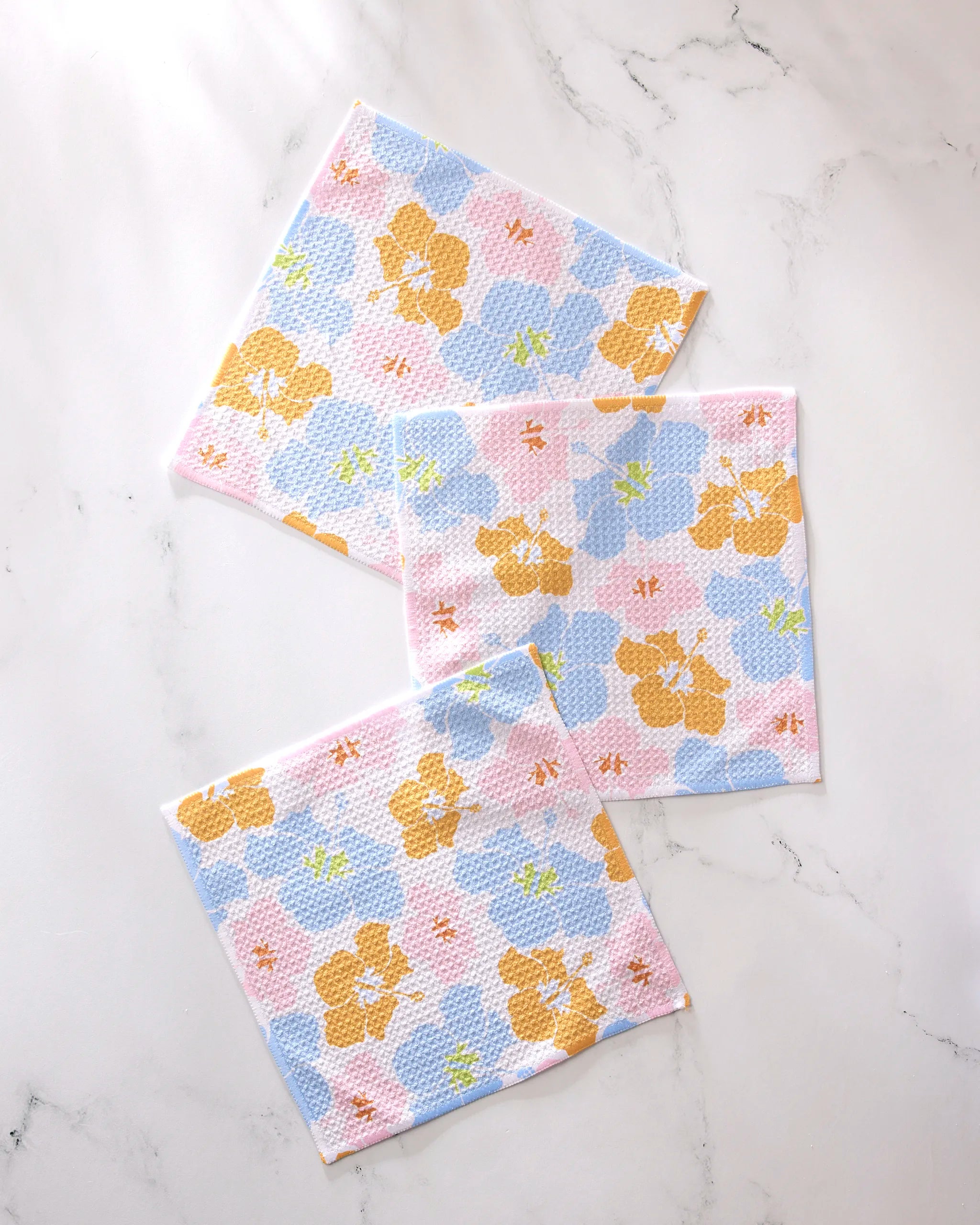 The GEOMETRY Hula Hues set features three super-absorbent square dishcloths with a blue, yellow, pink, and green floral pattern. Arranged overlapping on white marble, each cloth has a textured look.