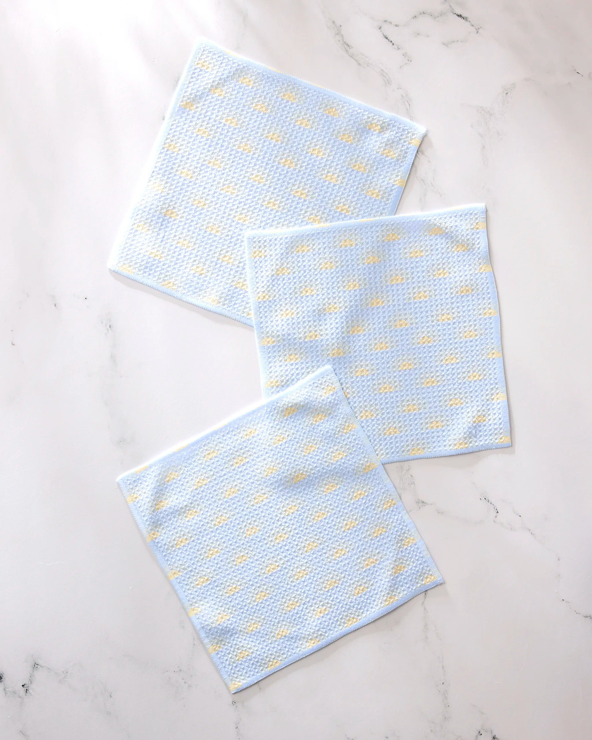 Three GEOMETRY "Soft Summer Sun" light blue textured recycled dishcloths with small yellow dots are arranged on a white marble surface.