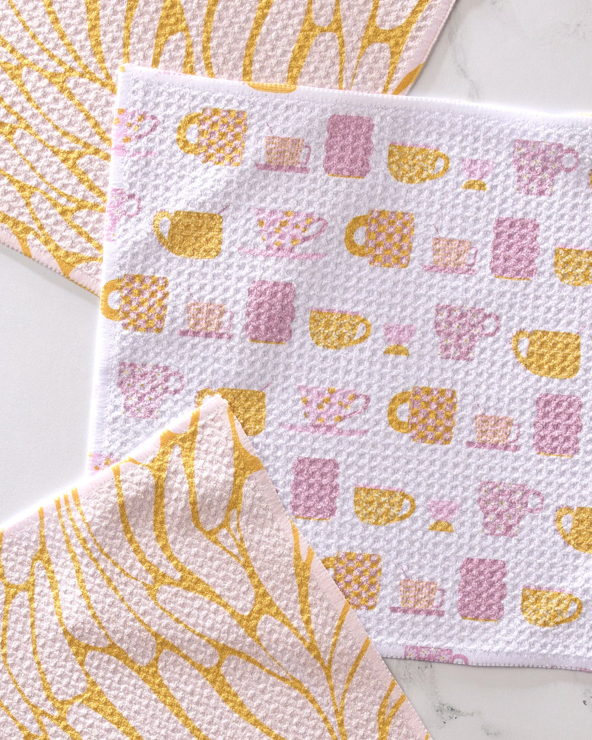Three Tenderhearted dishcloths by GEOMETRY, made from recycled materials, rest on a white surface: two have yellow and pale pink abstract leaf patterns, the center features pink and yellow mugs and cups.