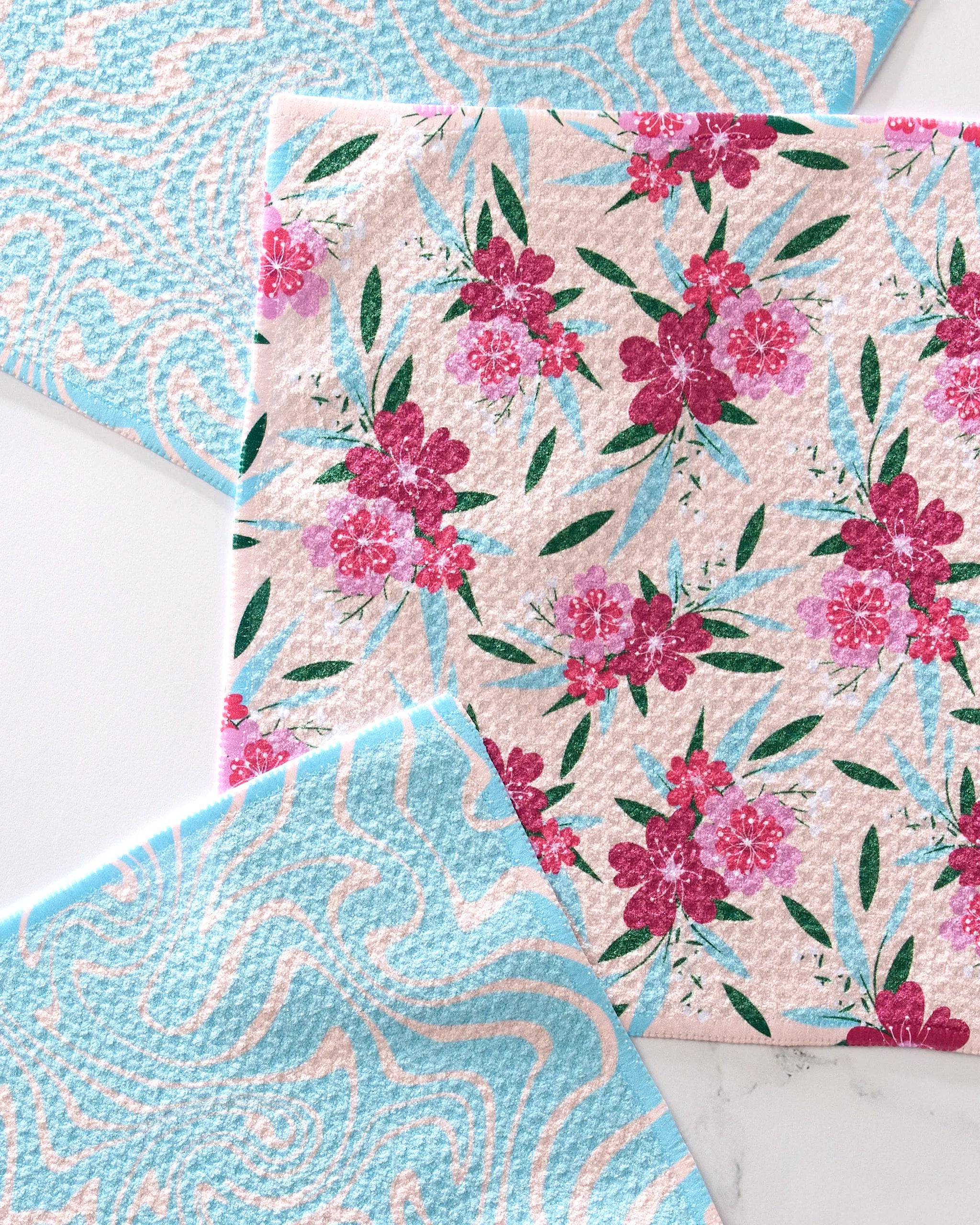 Three GEOMETRY "Sending Love" dishcloths rest on a white surface: two feature light blue and white wavy patterns, while the center displays a pink floral design with green leaves. Made from recycled materials for eco-friendly style.