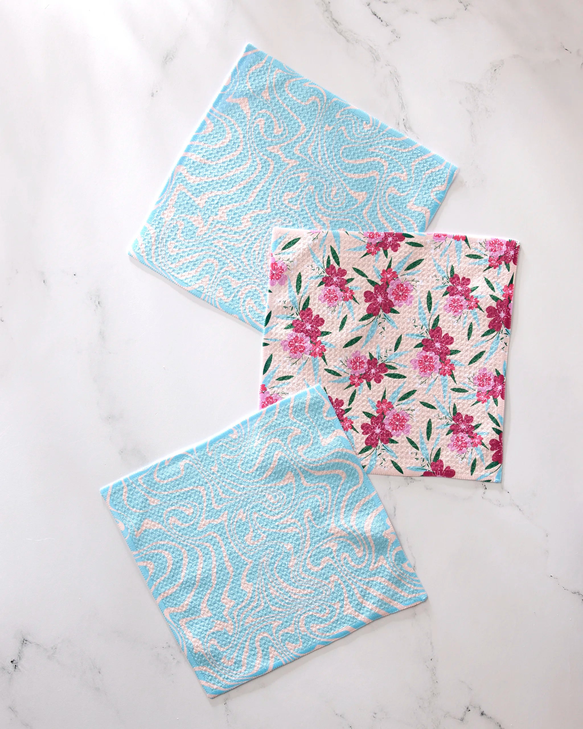 The GEOMETRY "Sending Love" set features three reusable cloth wipes made from recycled materials: two light blue with wavy patterns and one with a pink floral design and green leaves, all displayed on a white marble surface.
