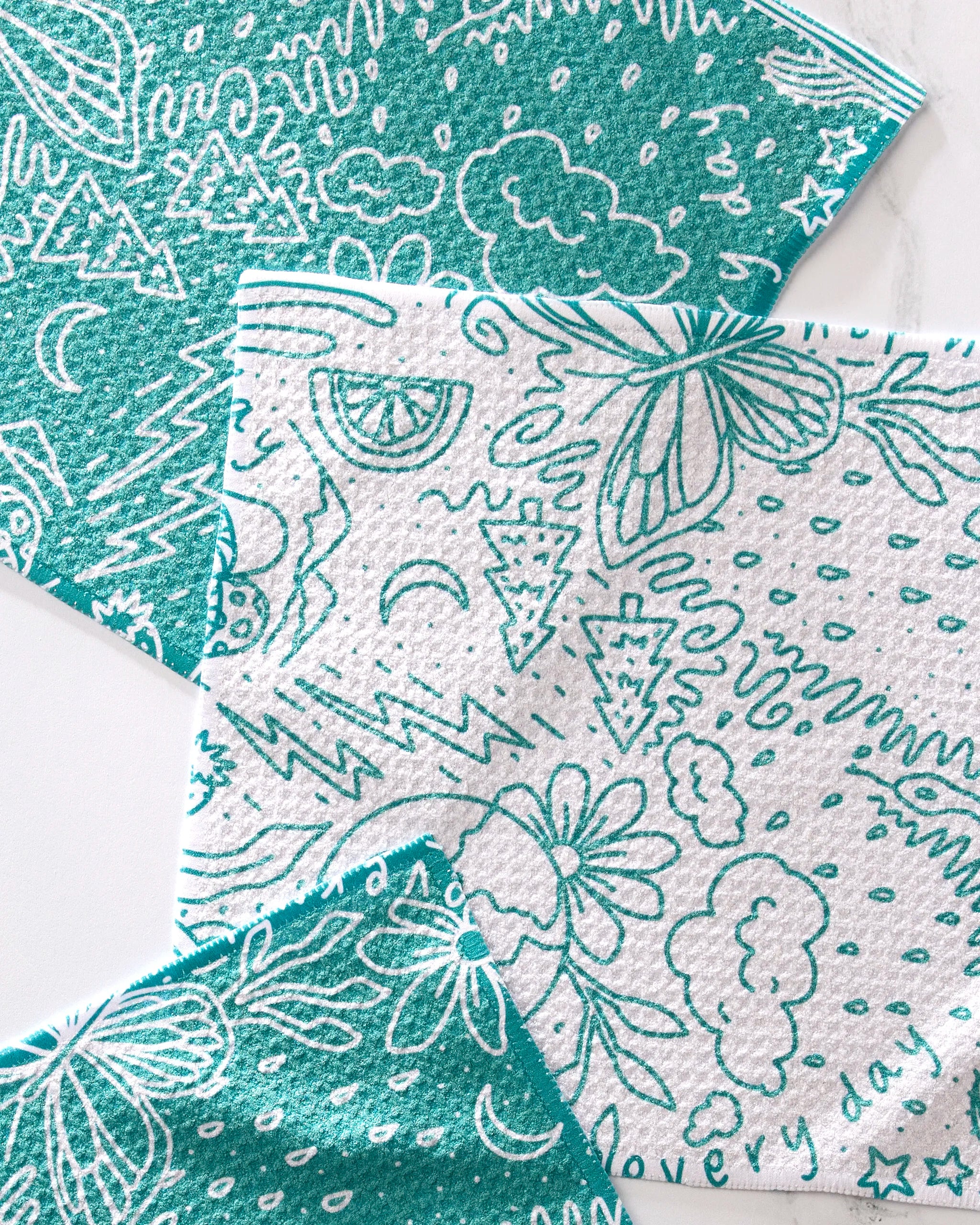 The GEOMETRY Every Day set includes three overlapping teal and white dishcloths featuring playful, doodle-inspired designs—flowers, trees, butterflies, stars, lemons, and lightning bolts—on a white surface. Made from recycled materials.