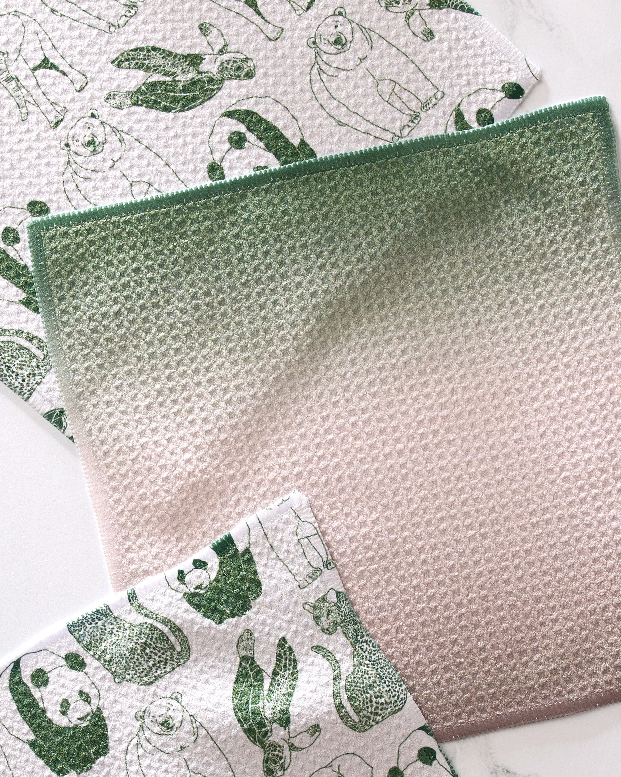 Three "Endangered" dishcloths by GEOMETRY rest on a white surface: two with green animal prints and one with a green-to-pink gradient. Made from recycled materials, these eco-friendly cloths are both super-absorbent and stylish.