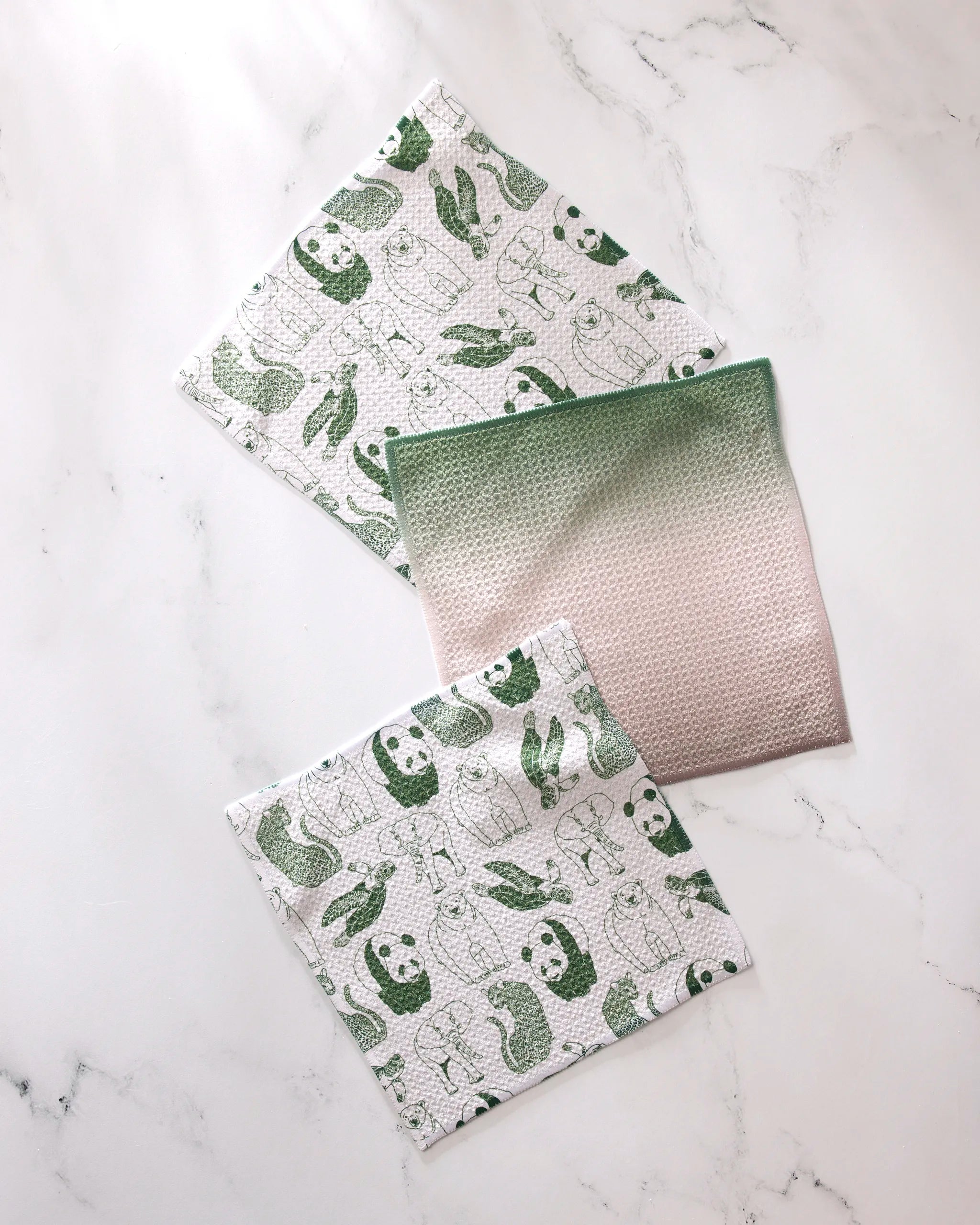 The GEOMETRY Endangered set features three reusable cloths: two with green animal prints (pandas, turtles, rabbits) and one pink cloth with a green gradient. Made from recycled materials for eco-friendly cleaning.