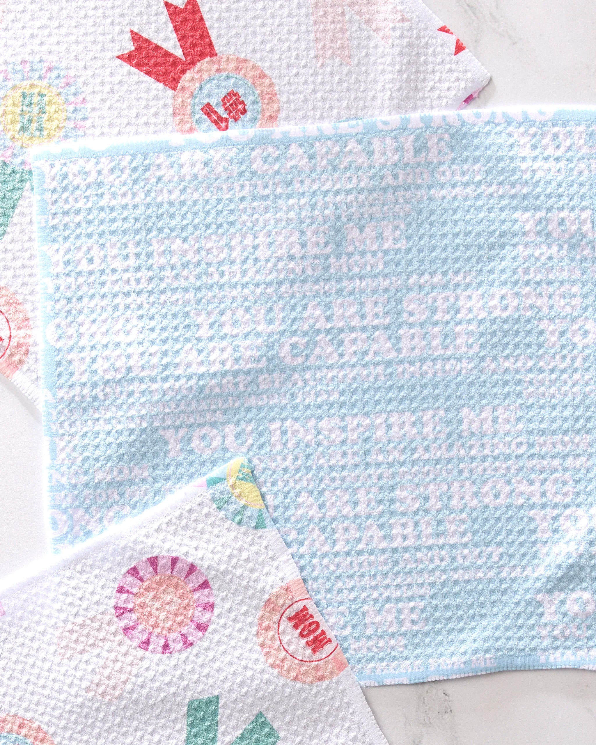 The GEOMETRY "Mom Mantra" dishtowel, blue and white with inspirational words, sits on marble with other recycled cloths featuring award ribbons and positive phrases.